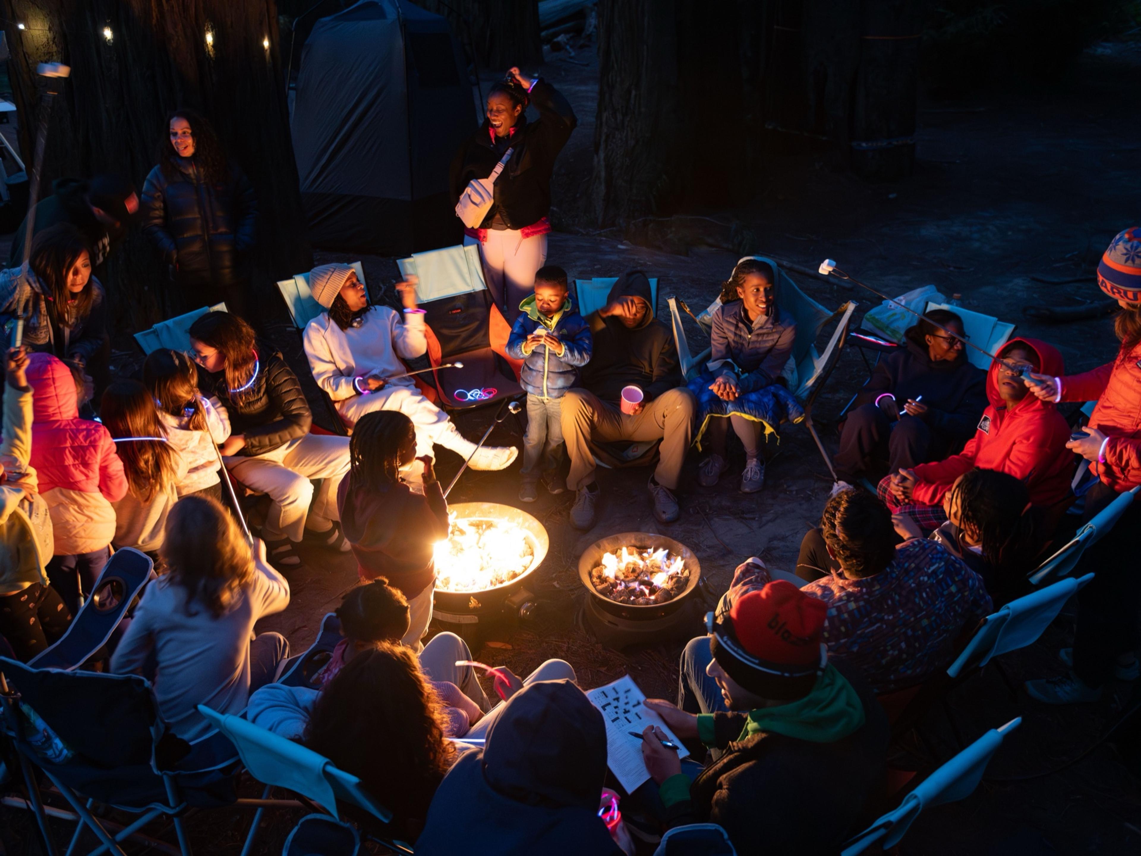 Night at a redwood campground: a circle of people around two propane fire pits, kids wearing glow-stick necklaces roasting marshmallows, string lights in the trees Night at a redwood campground: a circle of people around two propane fire pits, kids wearing glow-stick necklaces roasting marshmallows, string lights in the trees