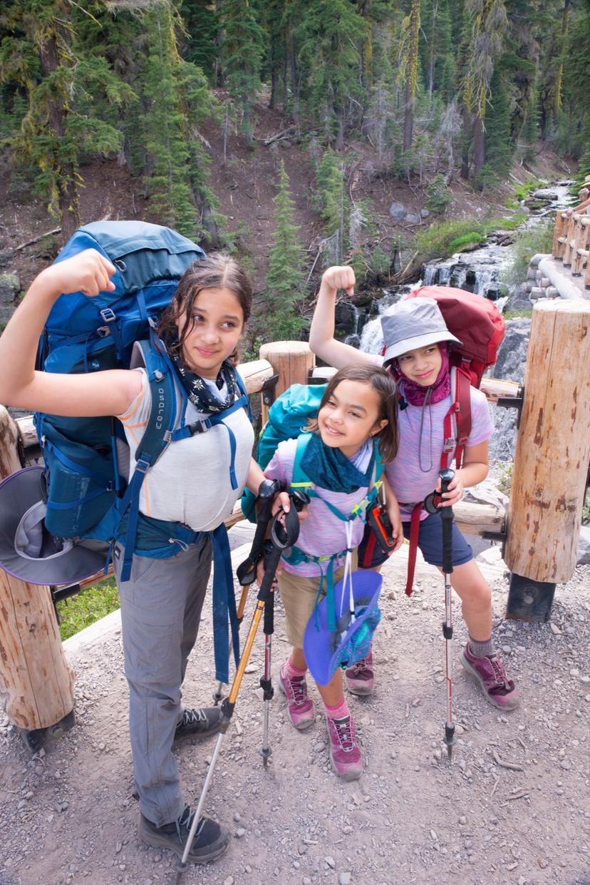 Three girls in sun hats with backpacking packs flexing on a footbridge along the Kings Creek trail in Lassen