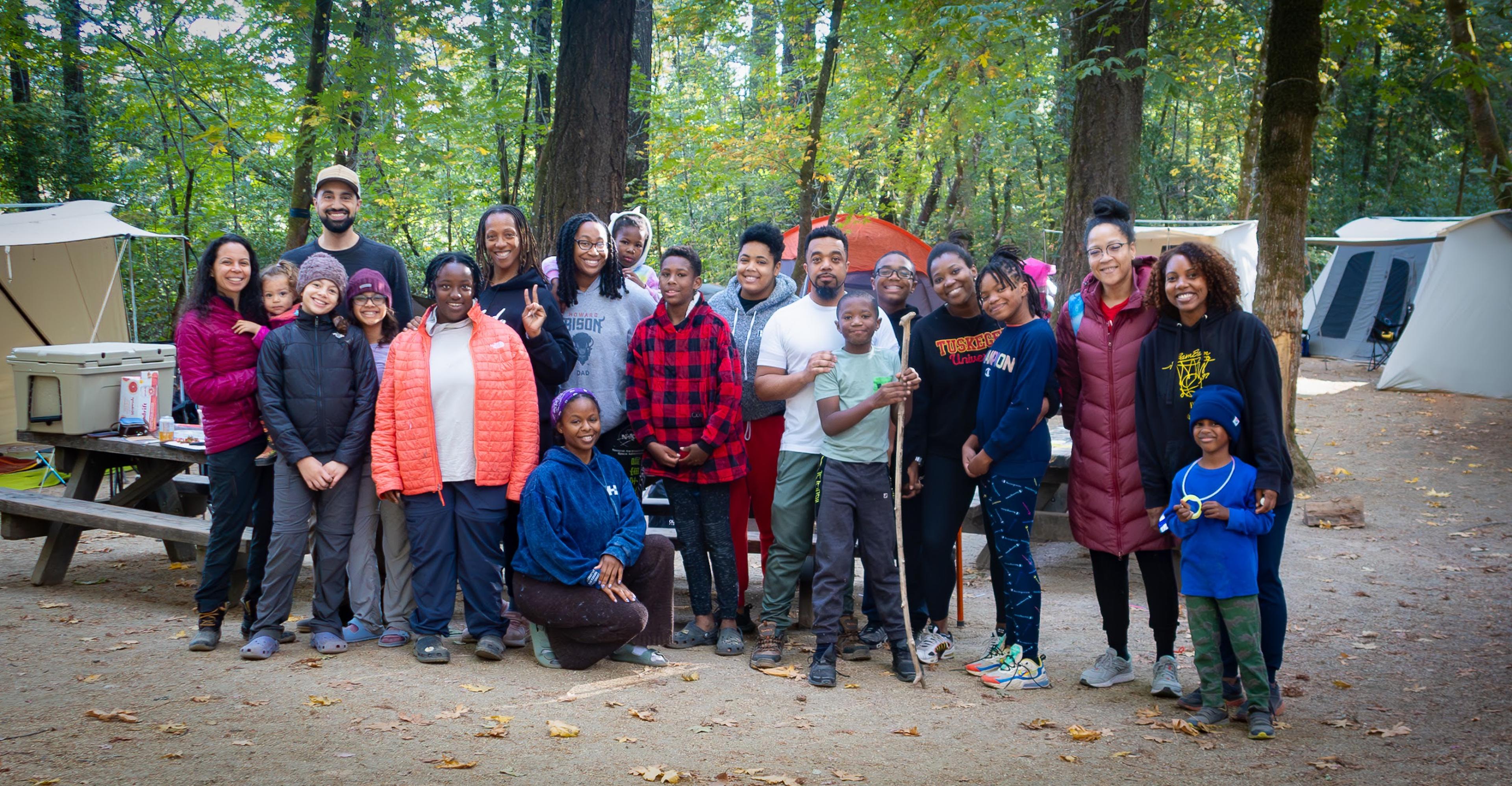 Diverse group of Outdoorithm Collective members of all ages standing together outdoors at a campsite, smiling and connected, representing what an inclusive outdoor community looks like in practice Diverse group of Outdoorithm Collective members of all ages standing together outdoors at a campsite, smiling and connected, representing what an inclusive outdoor community looks like in practice