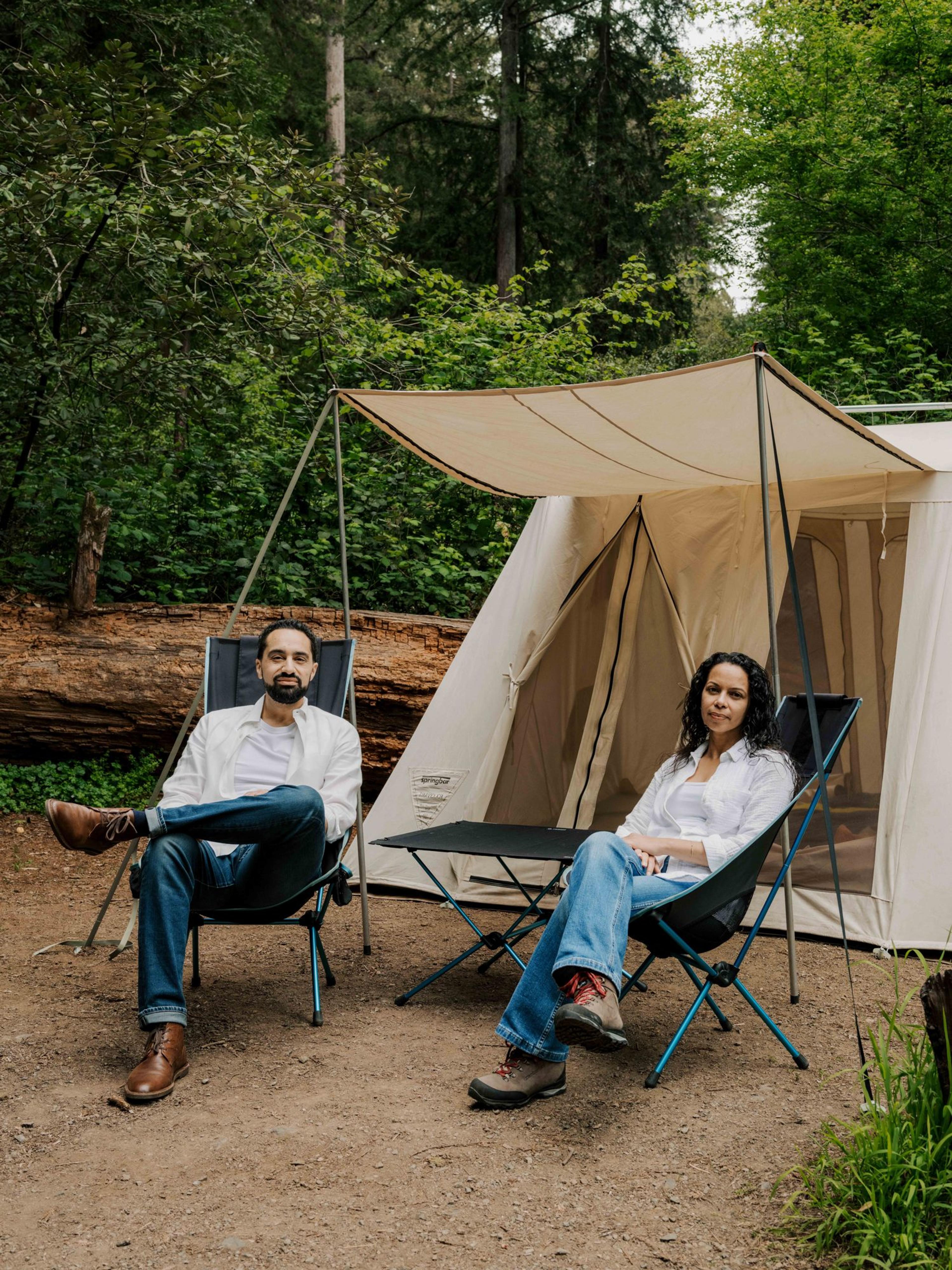 Two campers relaxing in high-back camp chairs outside a canvas tent with its awning up, redwoods behind Two campers relaxing in high-back camp chairs outside a canvas tent with its awning up, redwoods behind