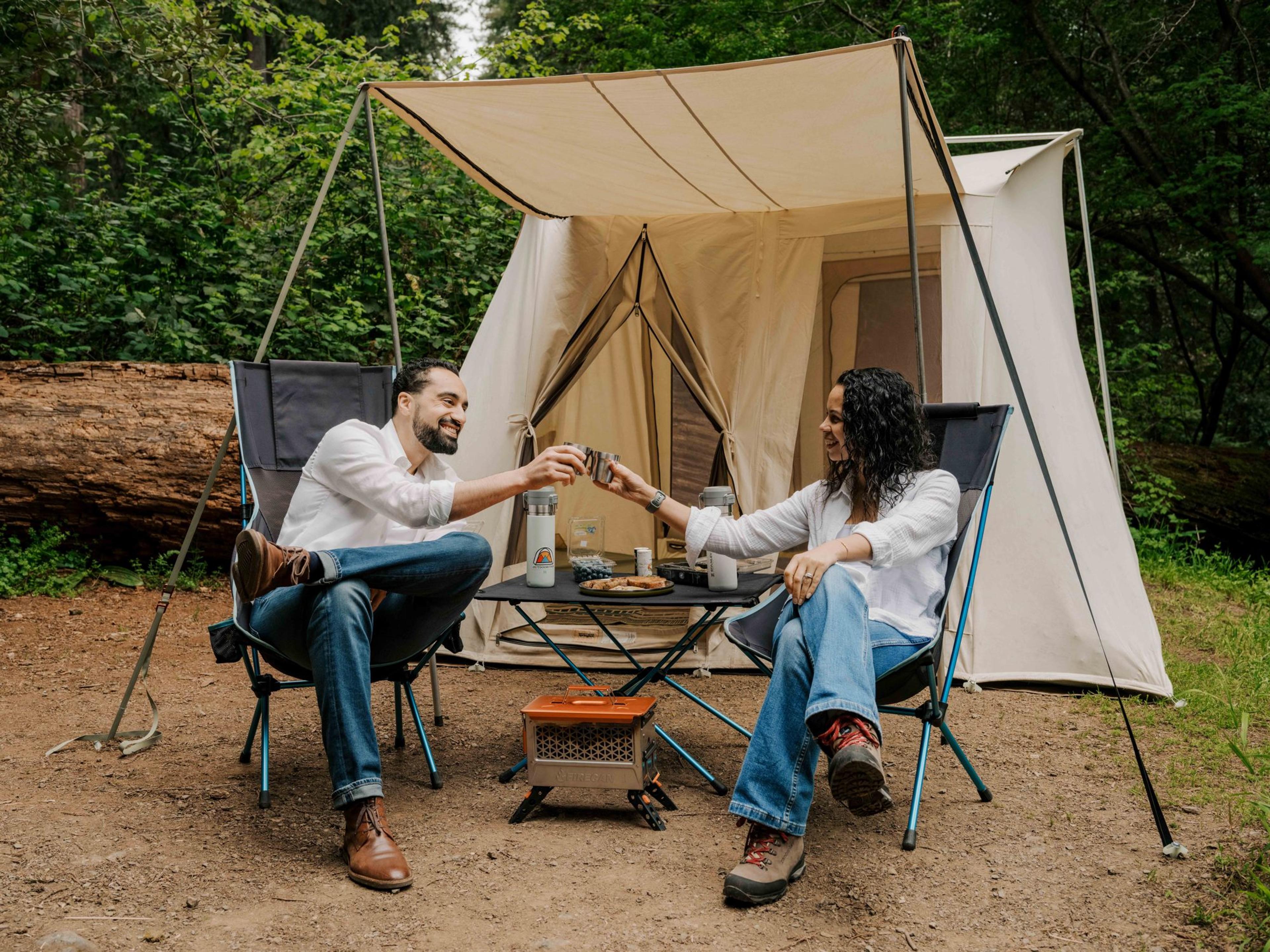 Two campers in high-back camp chairs clinking mugs across a low camp table, an orange Ignik FireCan between them and a canvas tent with its awning behind Two campers in high-back camp chairs clinking mugs across a low camp table, an orange Ignik FireCan between them and a canvas tent with its awning behind