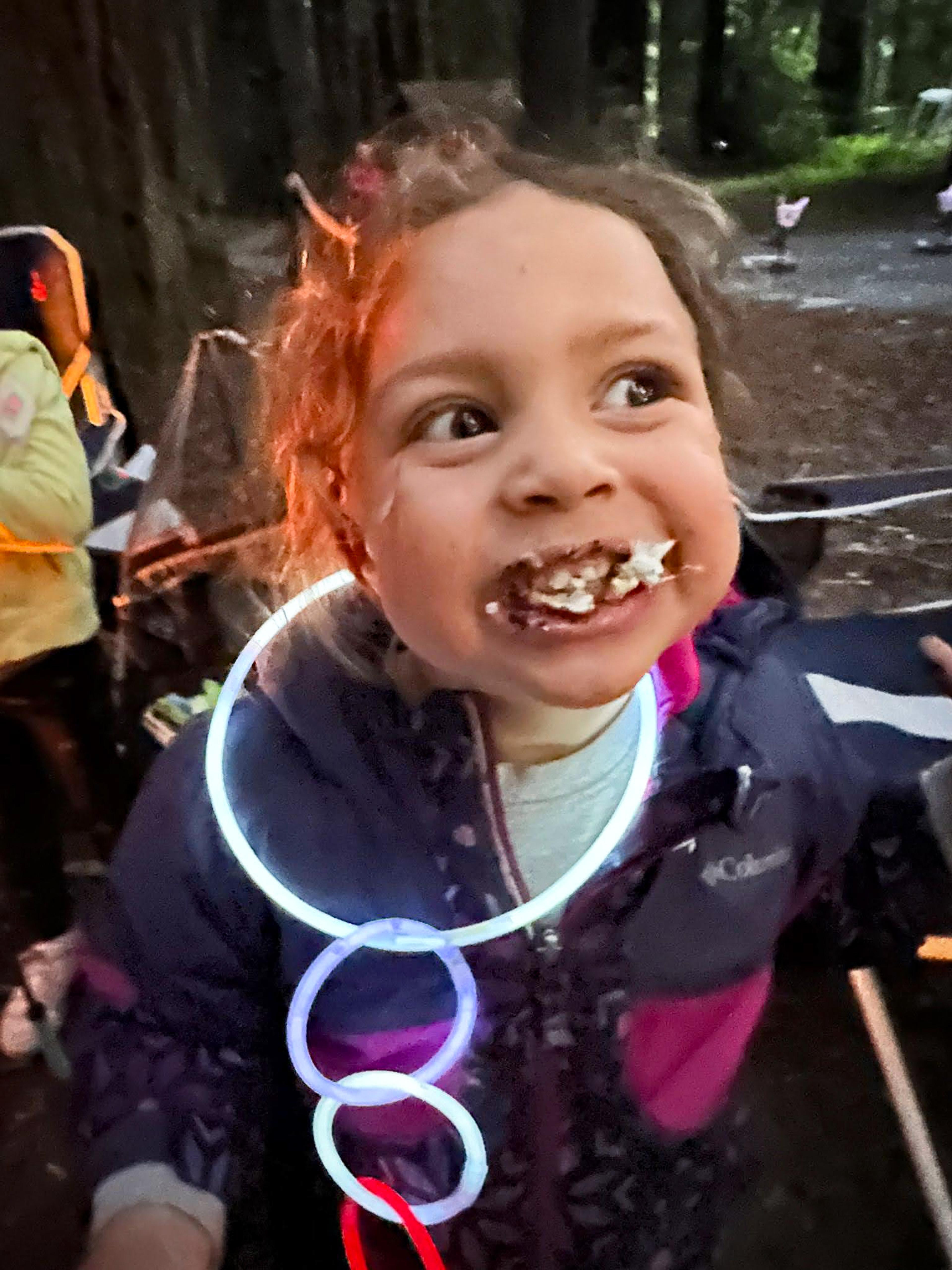 Close-up portrait of a young girl with curly hair and chocolate and marshmallow smeared around her mouth in a wide grin, wearing a purple Columbia jacket and a glowstick necklace, with forest trees and other campers blurred in the background at Williams Grove campground Close-up portrait of a young girl with curly hair and chocolate and marshmallow smeared around her mouth in a wide grin, wearing a purple Columbia jacket and a glowstick necklace, with forest trees and other campers blurred in the background at Williams Grove campground