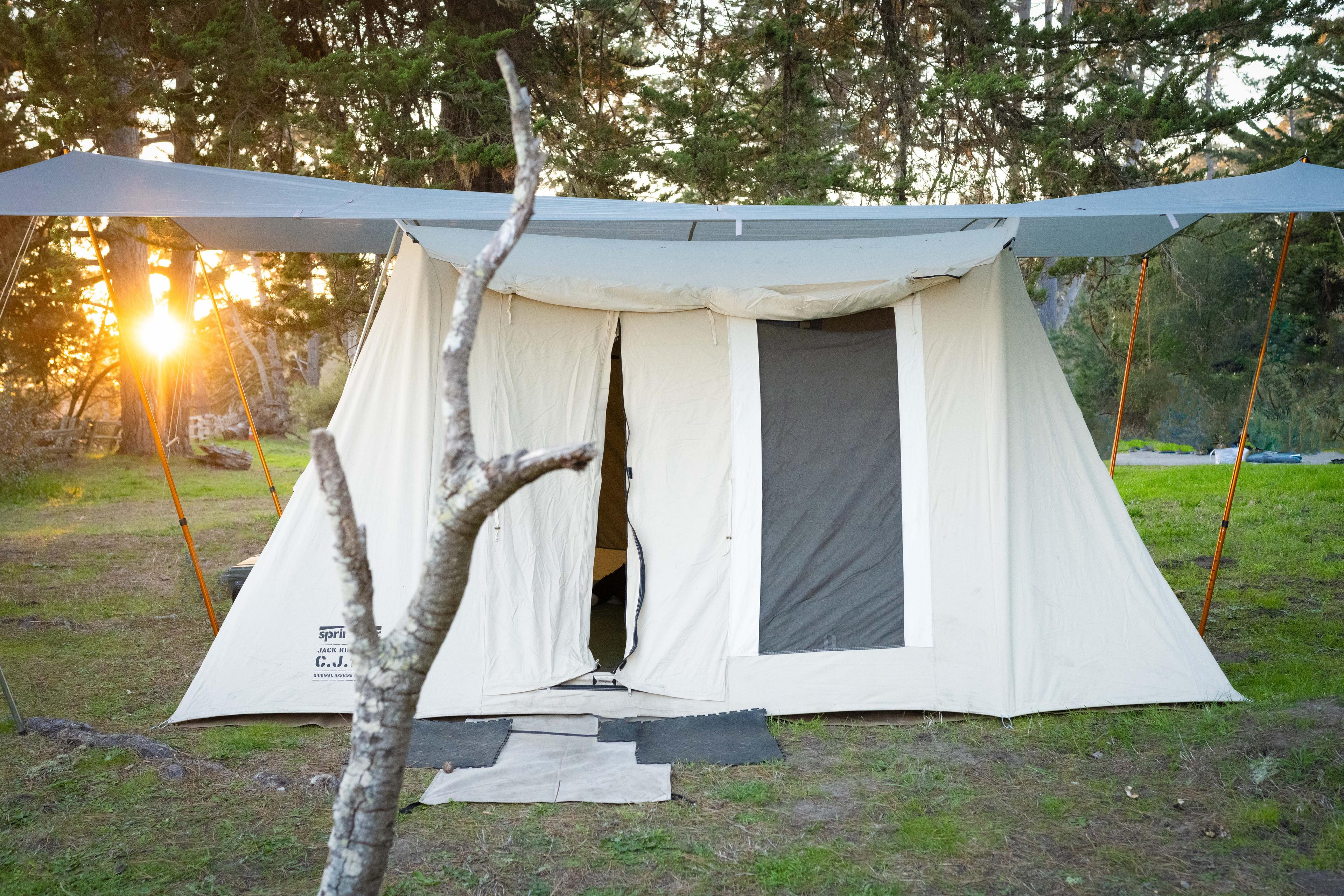 Large rectangular tarp pitched over a Springbar Classic Jack 140 canvas tent at a coastal campsite, anchored to nearby trees with paracord and trekking poles, designed to keep dew and rain off the canvas overnight