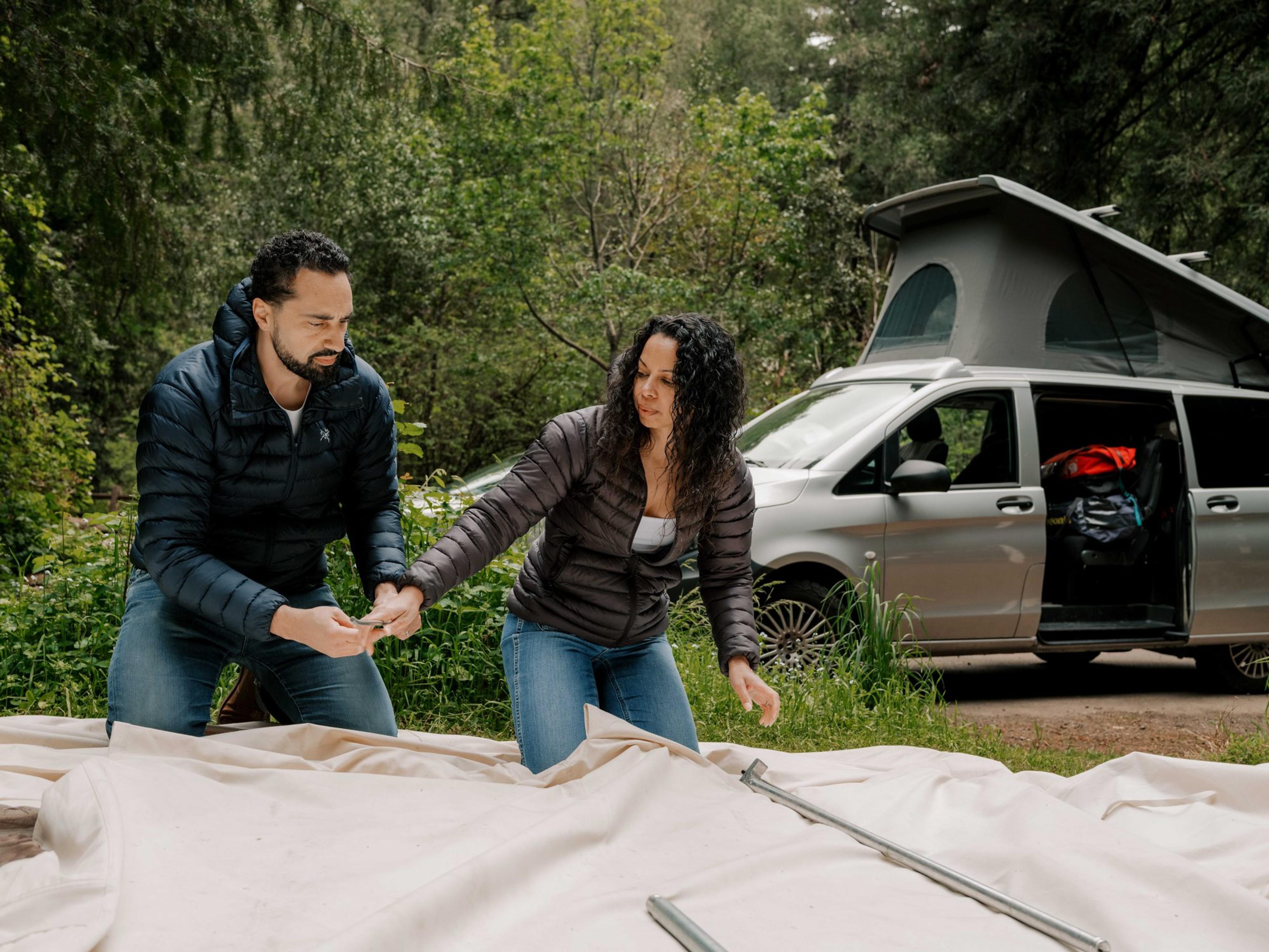 Two campers spreading out a canvas tent body on the ground with the poles laid on top, a camper van parked behind Two campers spreading out a canvas tent body on the ground with the poles laid on top, a camper van parked behind