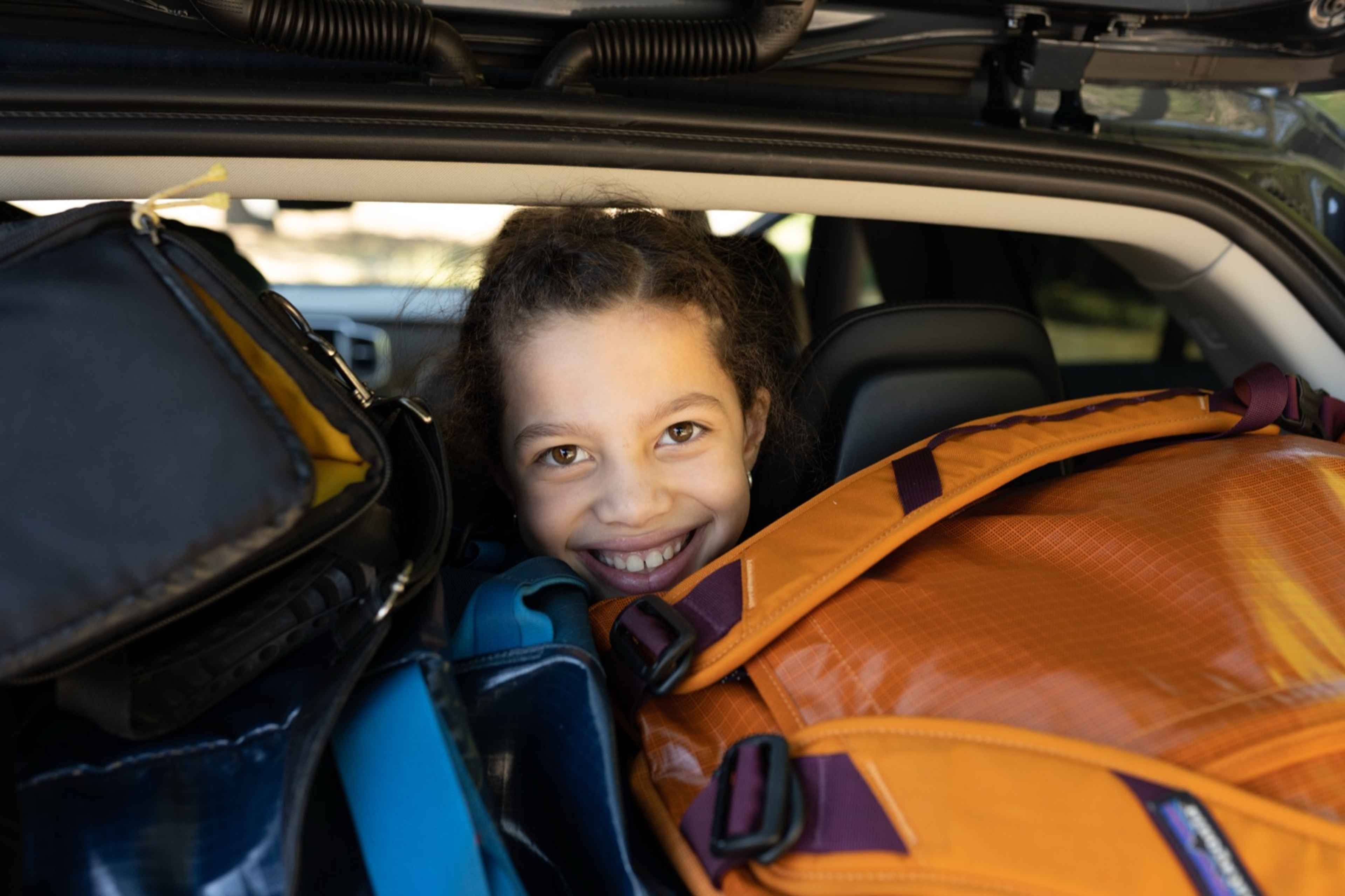 A smiling girl peeking out from the back of a car packed full of camping duffels and gear
