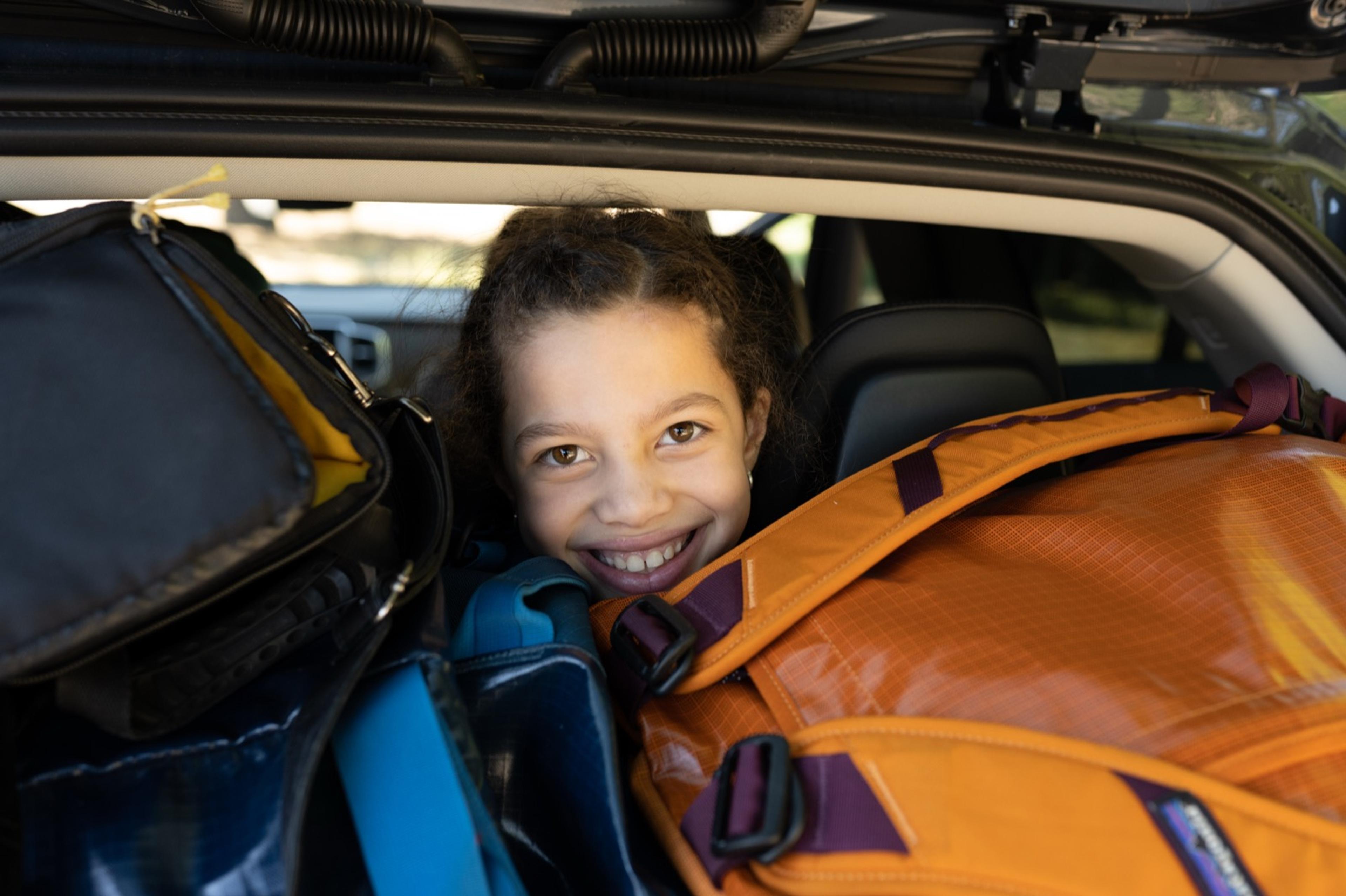 A smiling girl peeking out from the back of a car packed full of camping duffels and gear