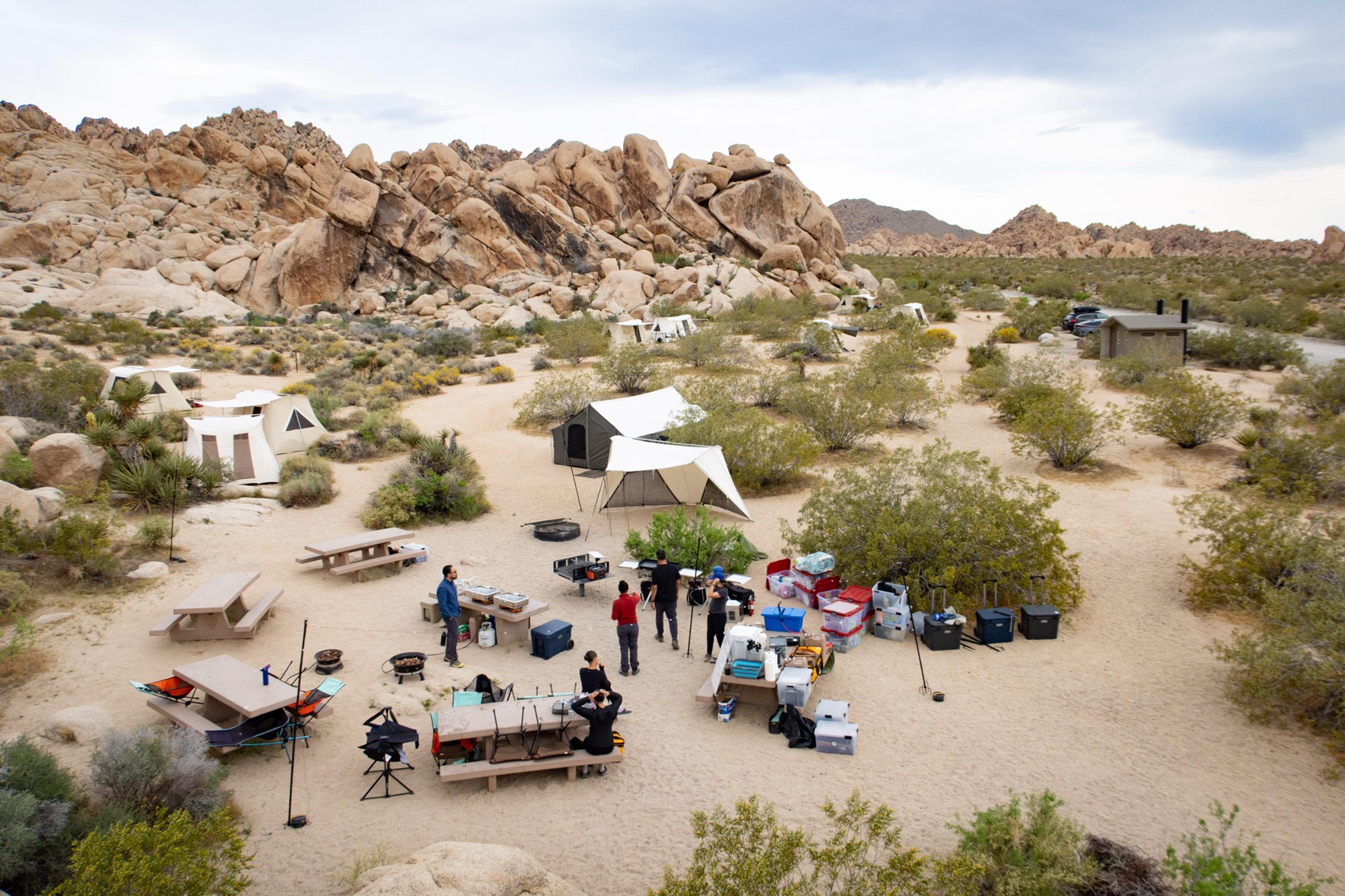 Aerial view of Indian Cove Group Campground at Joshua Tree showing Springbar canvas tents and a shared kitchen setup arranged across the desert sand surrounded by massive granite boulders