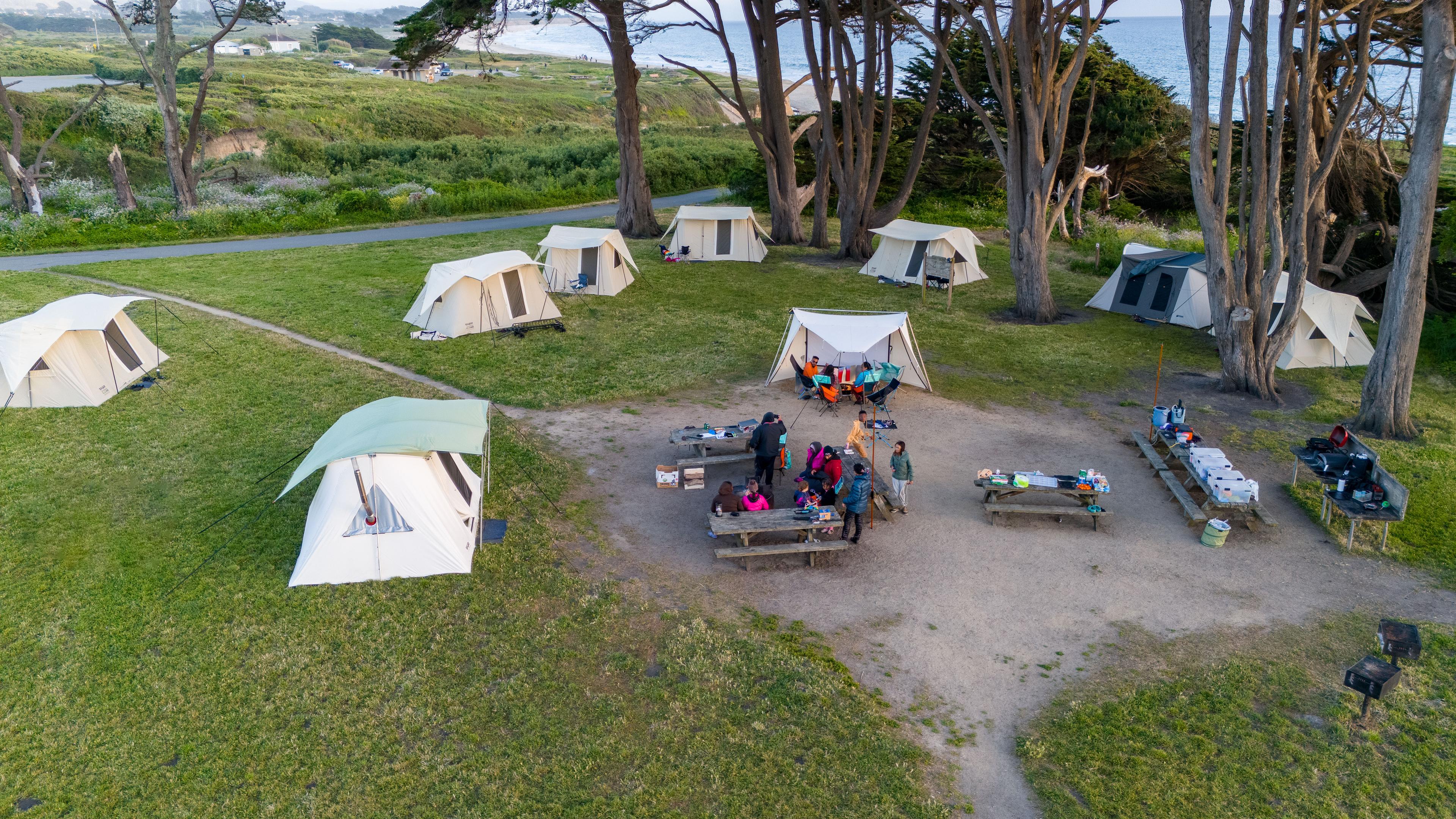 Aerial drone view of an Outdoorithm Collective group campsite at Half Moon Bay State Beach showing multiple Springbar canvas tents arranged around a shared central kitchen area with picnic tables, the Pacific Ocean visible in the background
