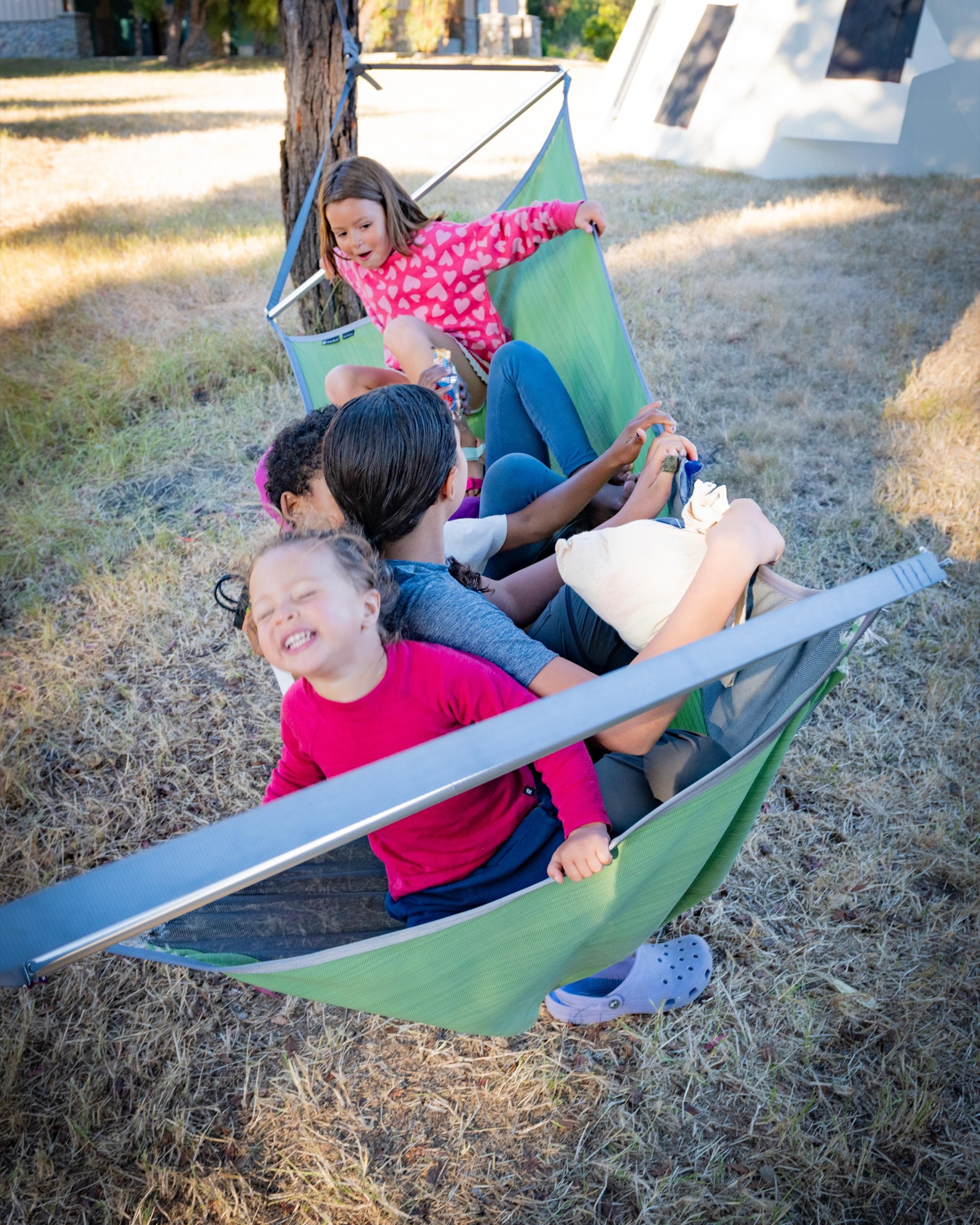 Several kids piled together laughing in a green spreader-bar hammock on a stand Several kids piled together laughing in a green spreader-bar hammock on a stand