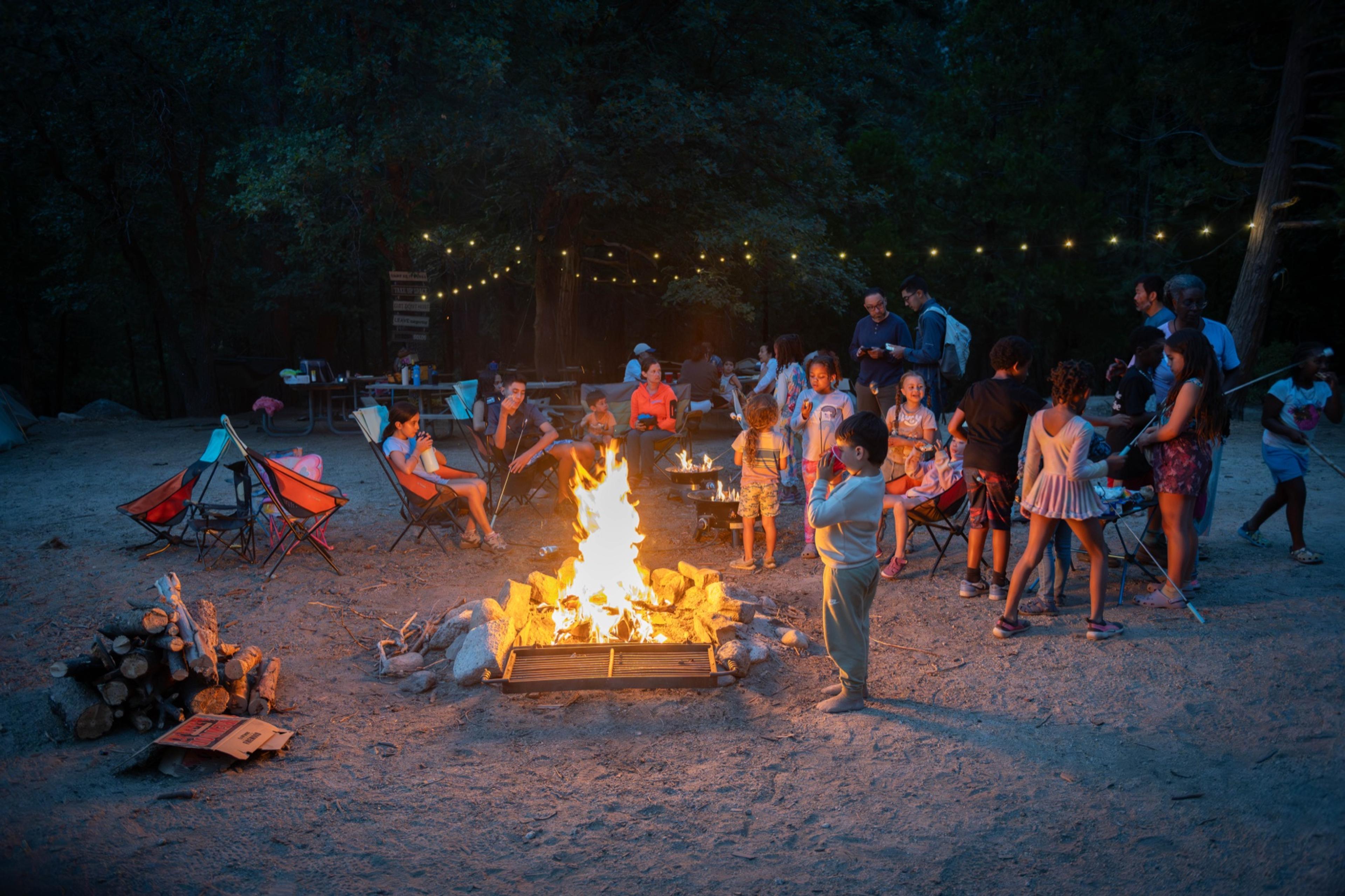 A large group gathered in camp chairs around a wood campfire at night in Kings Canyon, string lights overhead and a child standing near the flames A large group gathered in camp chairs around a wood campfire at night in Kings Canyon, string lights overhead and a child standing near the flames