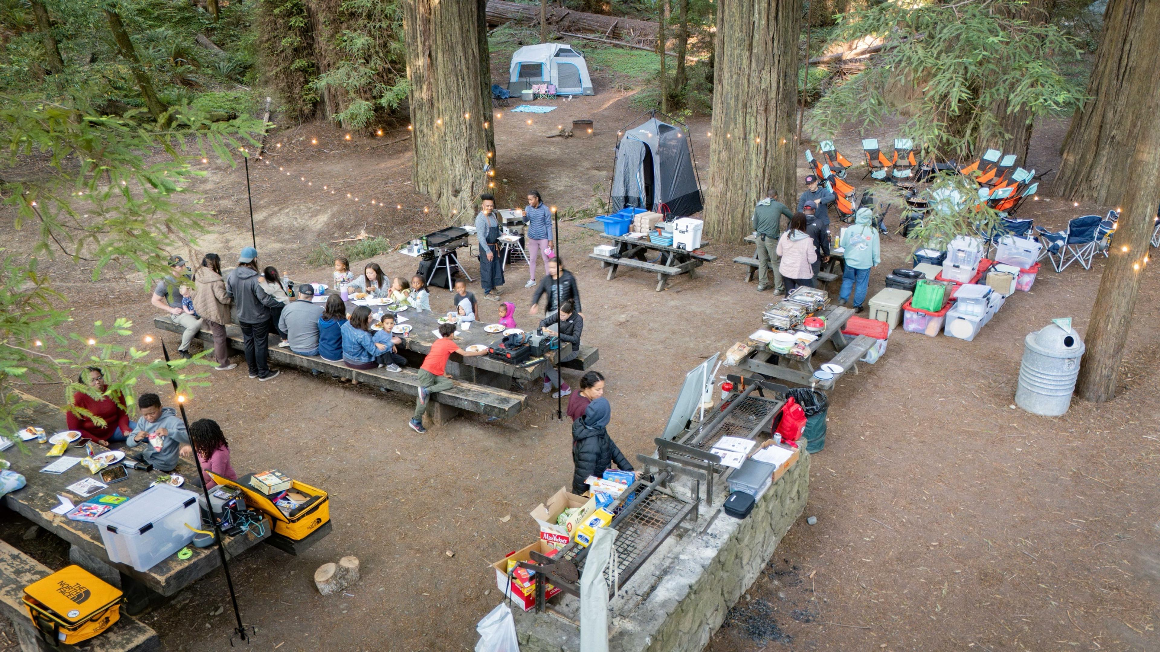 Overhead view of a group campsite in the redwoods: families at long picnic tables under string lights, a camp kitchen with coolers and storage bins, and a circle of camp chairs