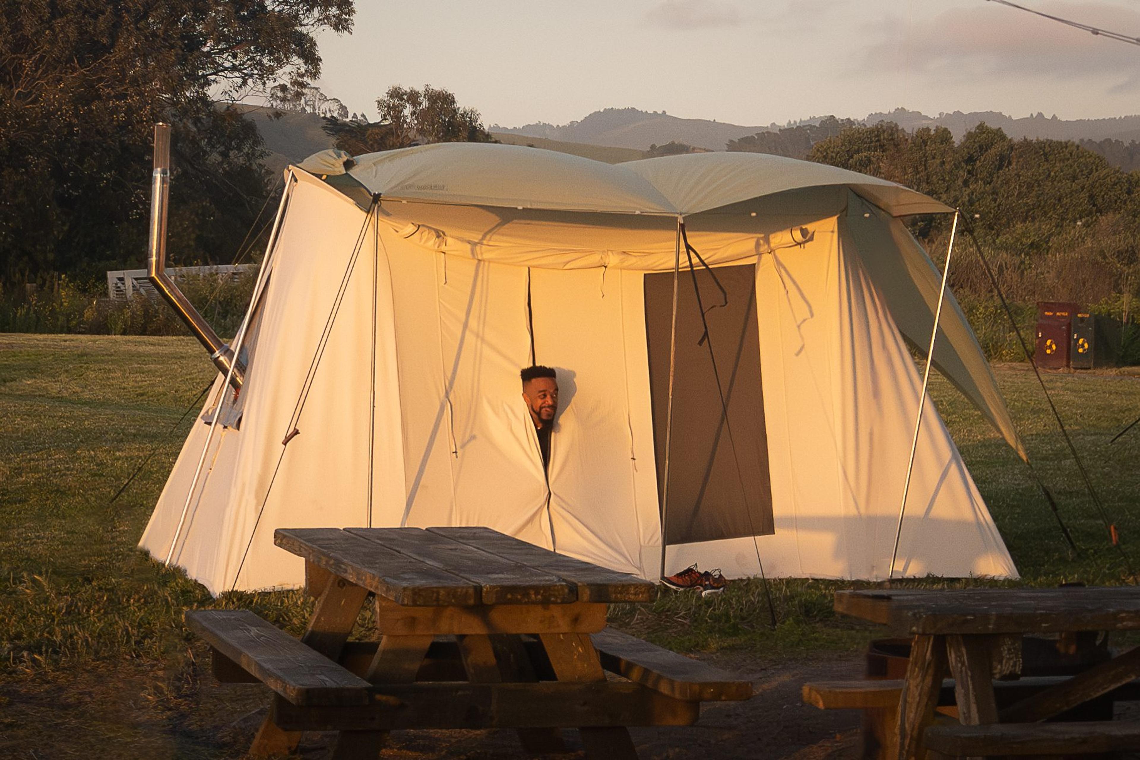 A white canvas wall tent with a stove pipe glowing at golden hour, a light on inside, picnic tables in front A white canvas wall tent with a stove pipe glowing at golden hour, a light on inside, picnic tables in front