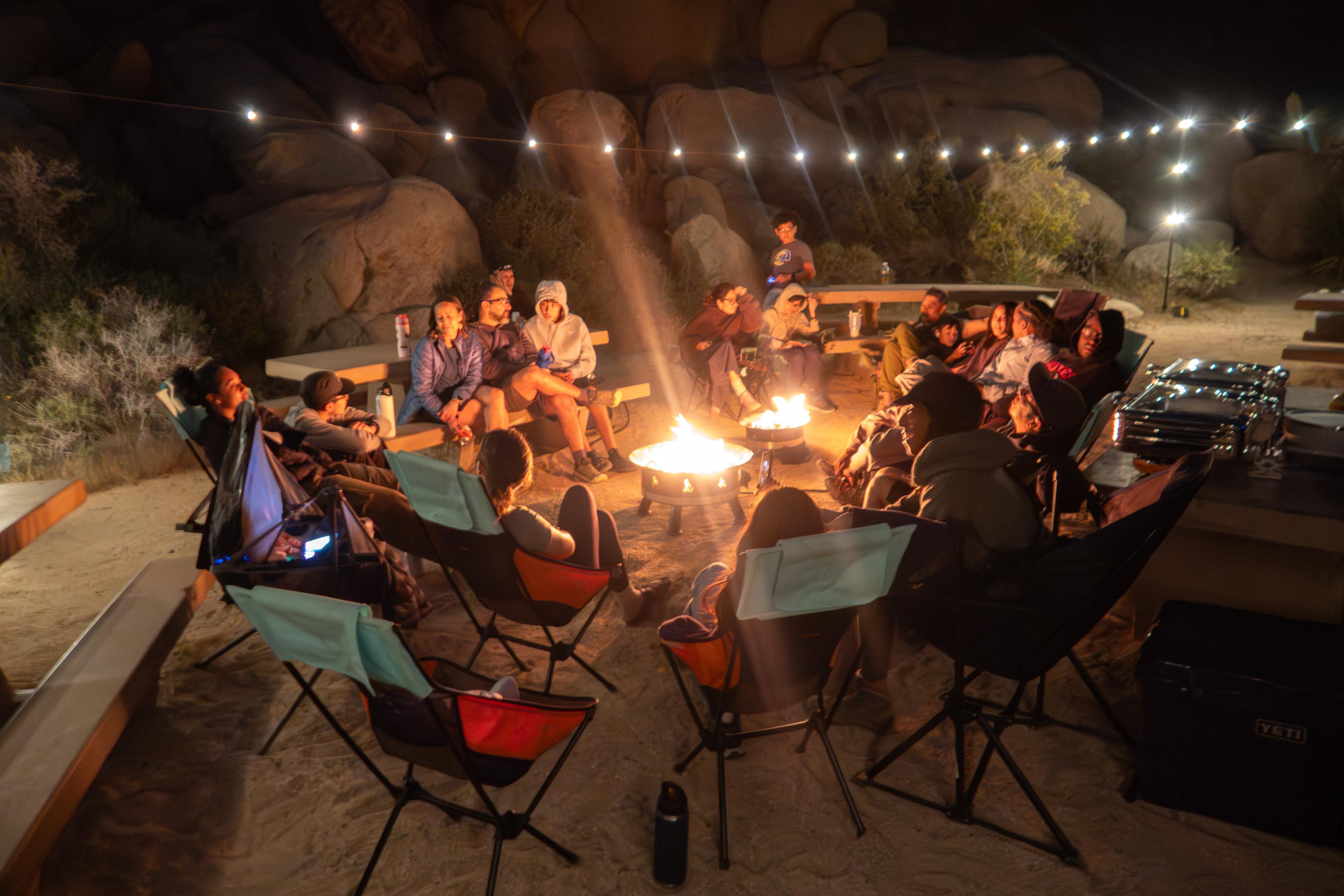 Twenty people in folding chairs in a circle around two propane fire pits at Indian Cove group campsite in Joshua Tree National Park at night, with string lights overhead and massive granite boulders in the background Twenty people in folding chairs in a circle around two propane fire pits at Indian Cove group campsite in Joshua Tree National Park at night, with string lights overhead and massive granite boulders in the background