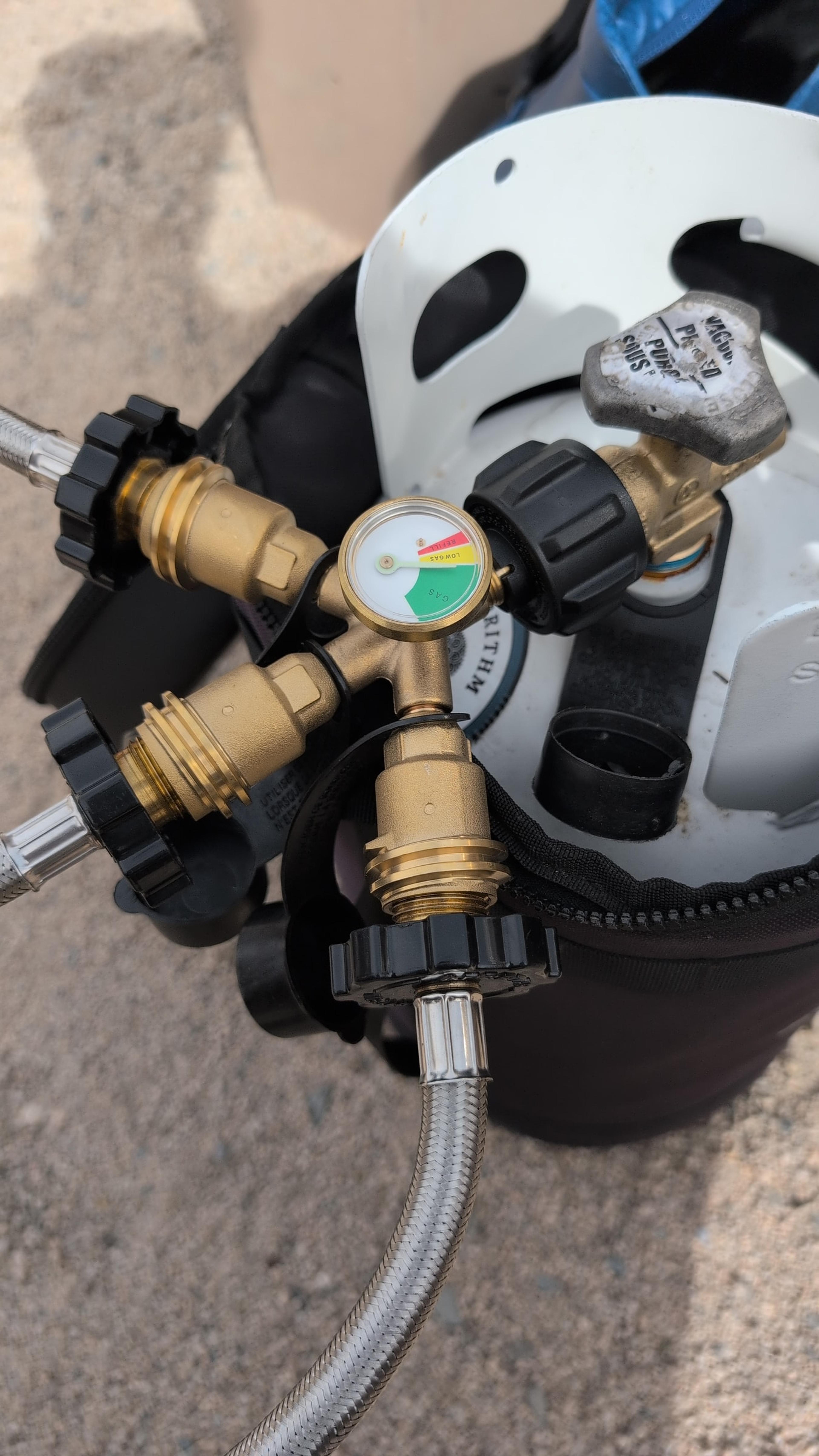 Brass triple propane adapter with pressure gauge mounted on a 10-pound propane tank in a black carry case Brass triple propane adapter with pressure gauge mounted on a 10-pound propane tank in a black carry case