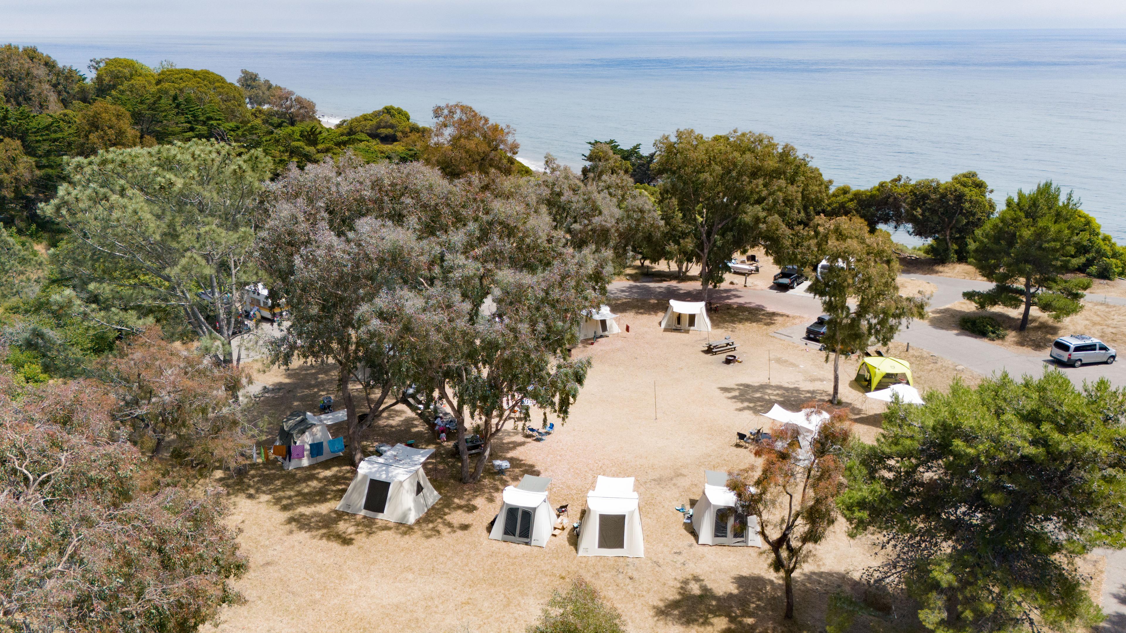 Aerial drone view of El Capitan State Beach campsite showing multiple white Springbar canvas tents arranged in a ring under a grove of oaks with the Pacific Ocean visible in the distance