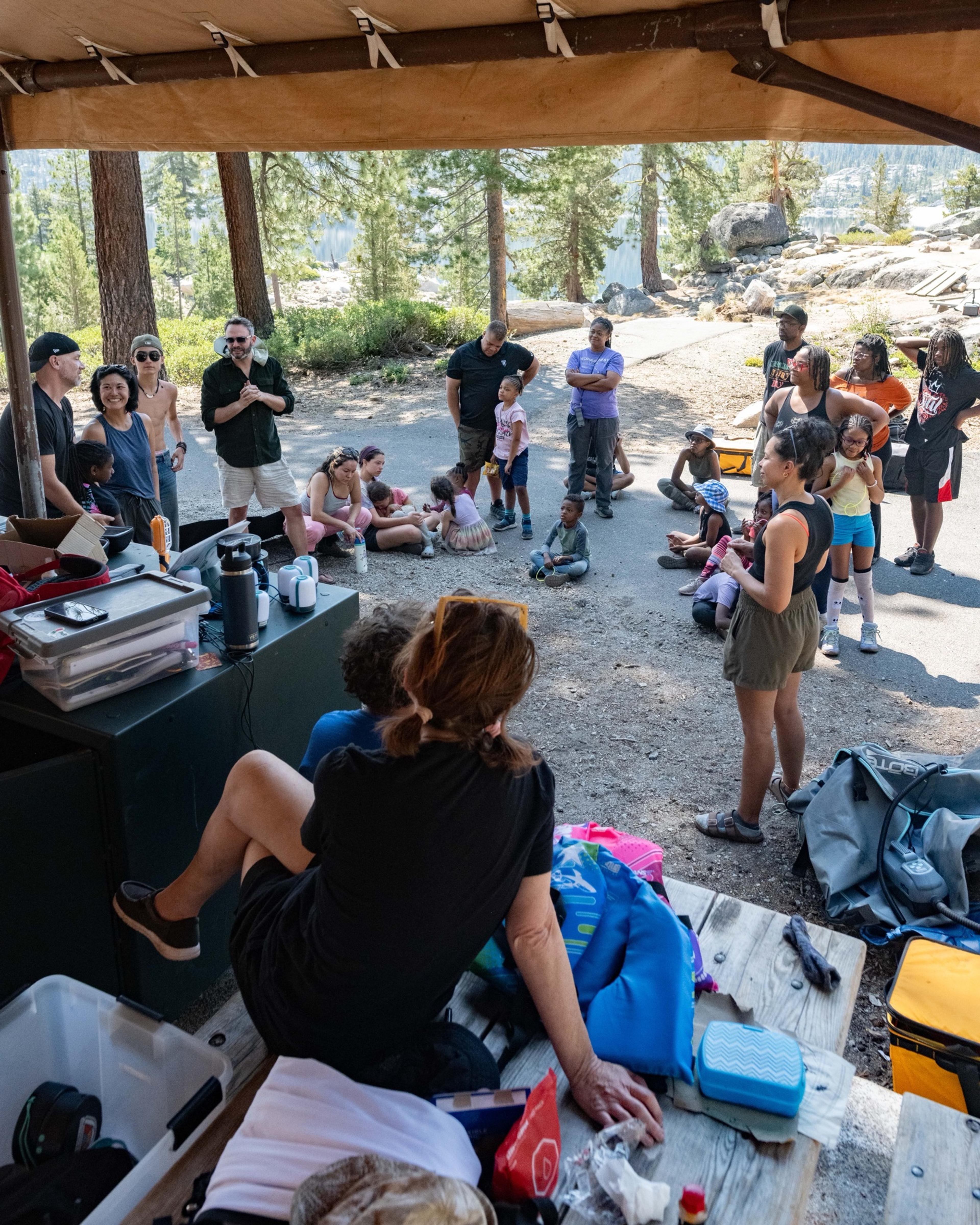 Group of about forty adults and children gathered under a canvas shelter at the Spicer Reservoir group campground for a morning meeting