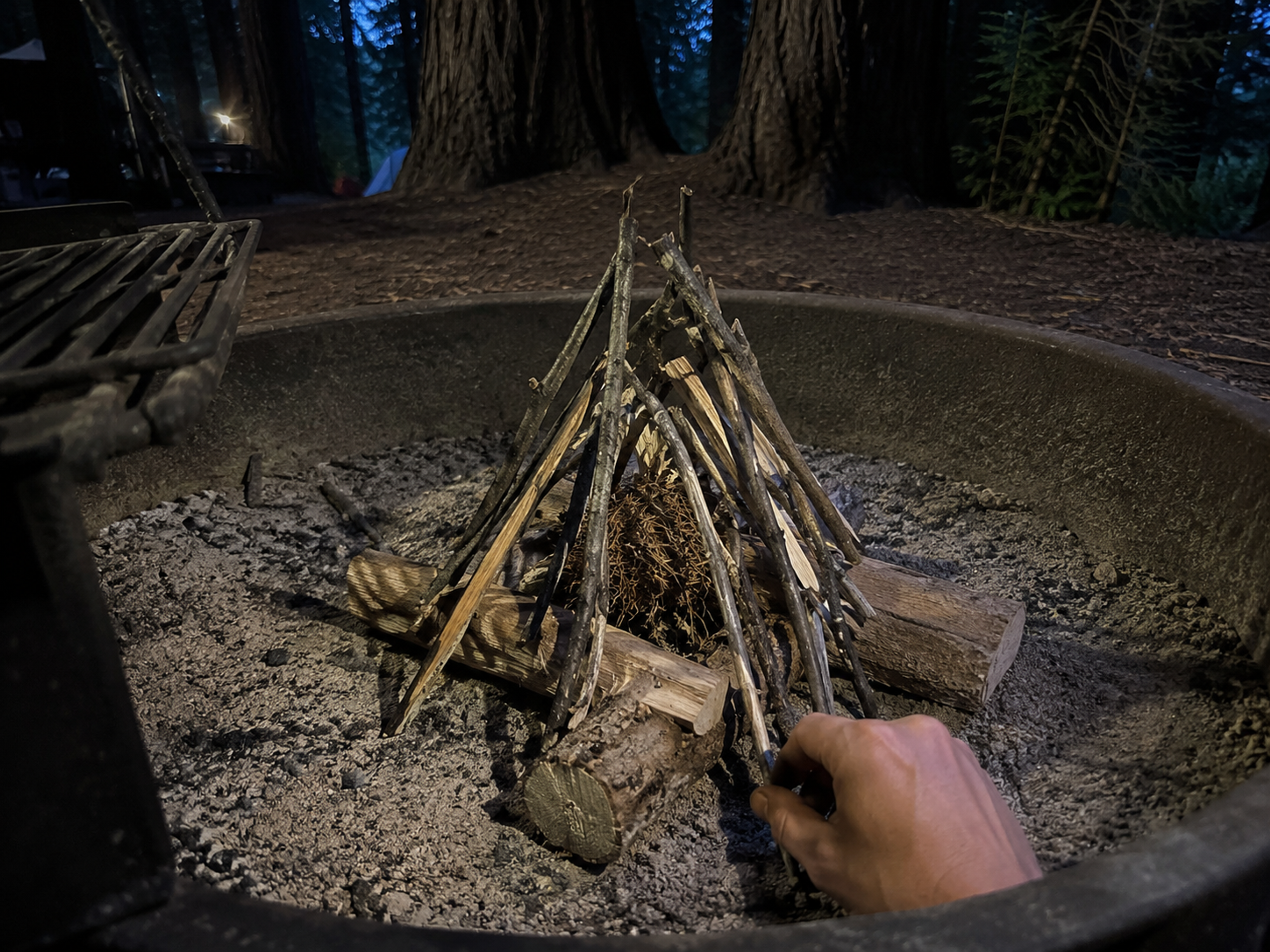 An unlit teepee-style campfire structure inside a metal fire ring at dusk: kindling sticks leaning inward in a cone shape around a center of tinder, with visible air gaps between sticks, a hand reaching in from the right placing another stick, redwood trees in soft background blur An unlit teepee-style campfire structure inside a metal fire ring at dusk: kindling sticks leaning inward in a cone shape around a center of tinder, with visible air gaps between sticks, a hand reaching in from the right placing another stick, redwood trees in soft background blur