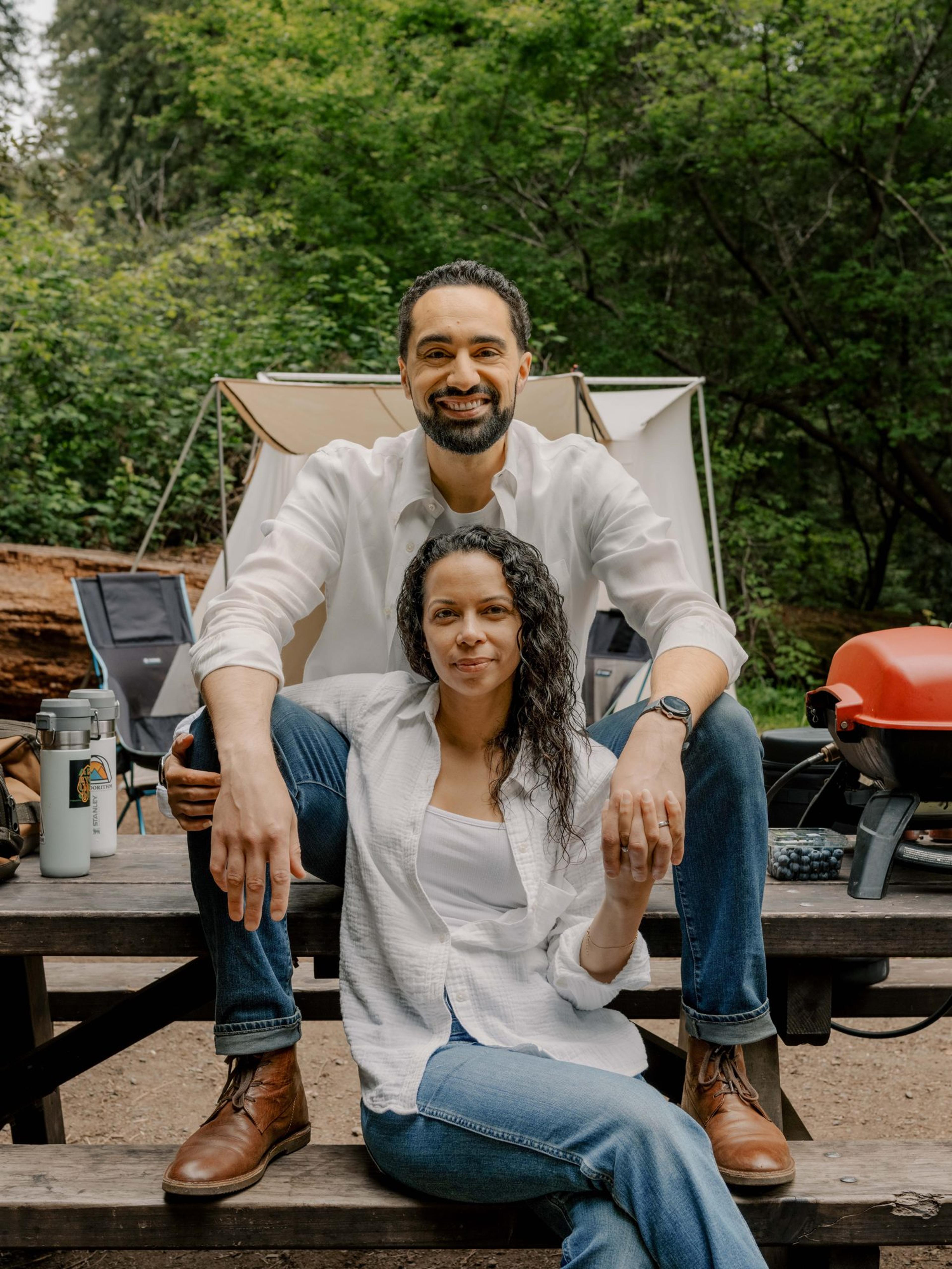 Two campers sitting on a picnic table in the woods beside a small red portable grill, a canvas tent behind them Two campers sitting on a picnic table in the woods beside a small red portable grill, a canvas tent behind them