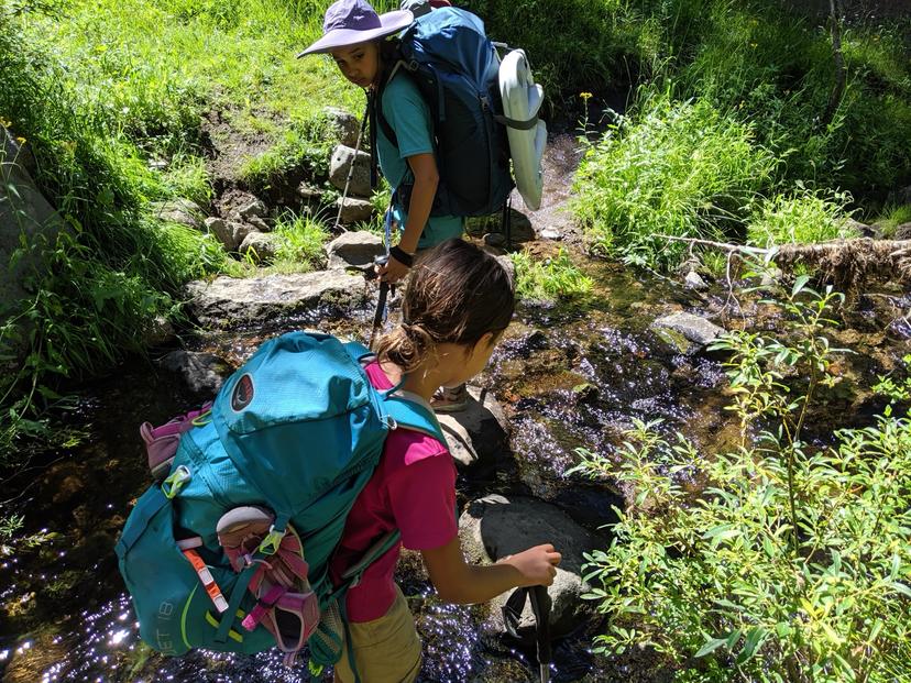 Two young girls wearing backpacking packs crossing rocks over Kings Creek surrounded by green brush in Lassen