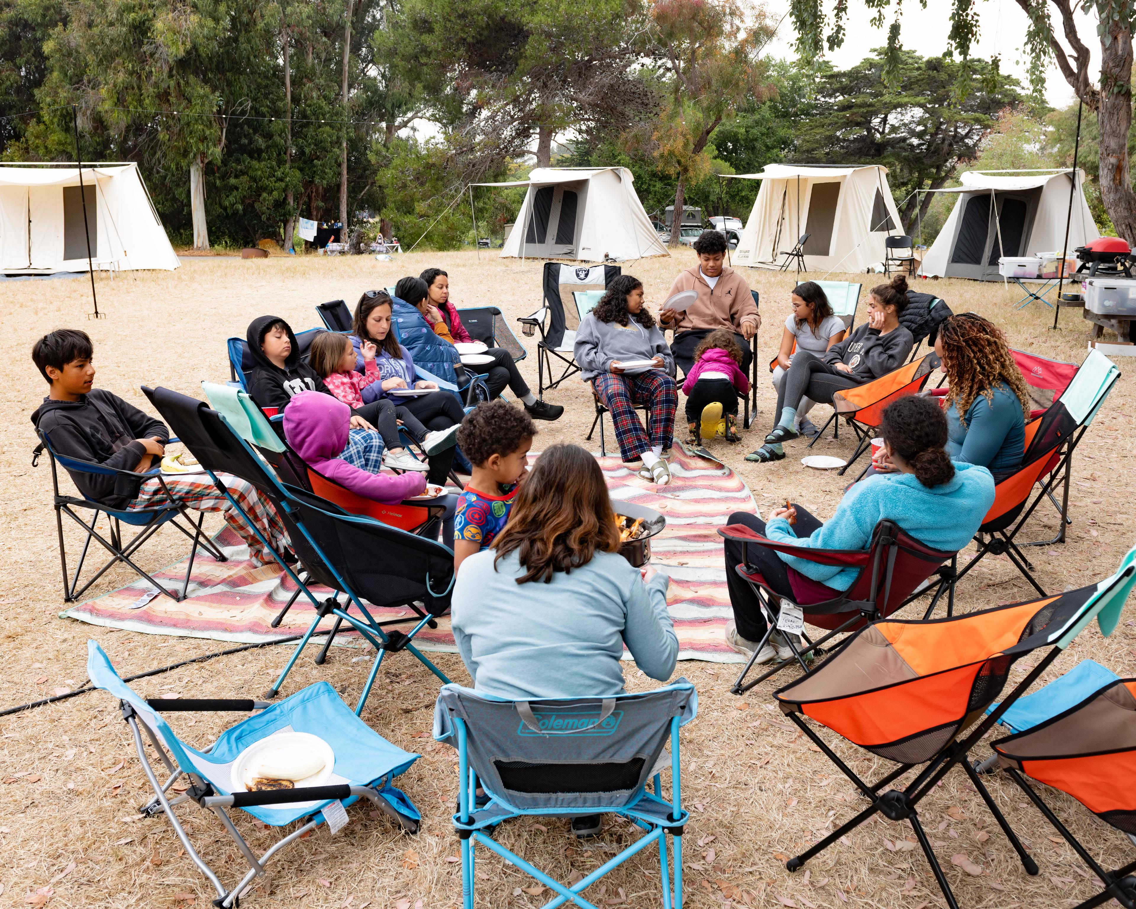 Outdoorithm Collective group of about 15 people seated in folding camp chairs in a circle on a Pendleton-style rug, with multiple white Springbar canvas tents pitched in the background at El Capitan State Beach group campground