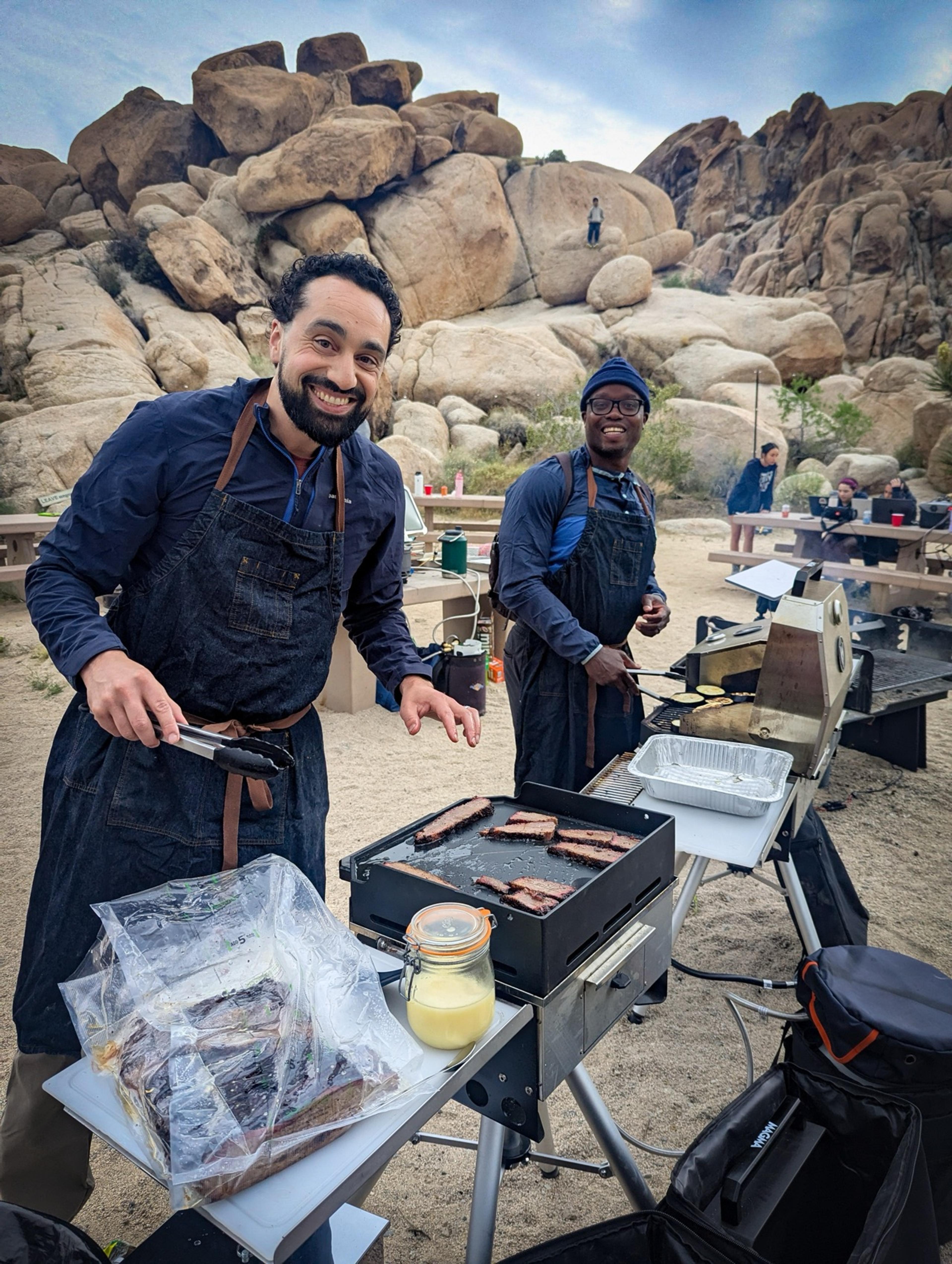 Two camp cooks working over a grill in the Joshua Tree desert at Indian Cove Group Campground, preparing brisket and ribs for the group