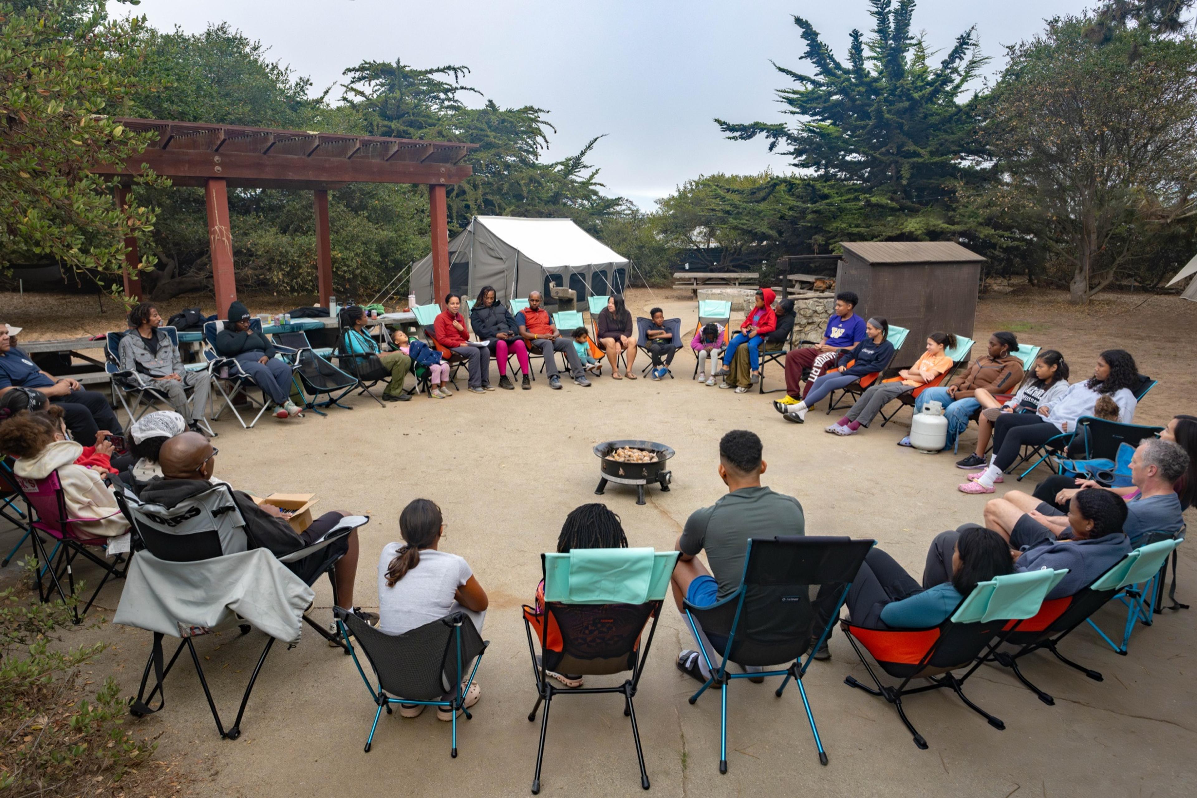 A large circle of high-back camp chairs around a portable propane fire bowl on an overcast afternoon, a canvas tent and cypress trees behind A large circle of high-back camp chairs around a portable propane fire bowl on an overcast afternoon, a canvas tent and cypress trees behind