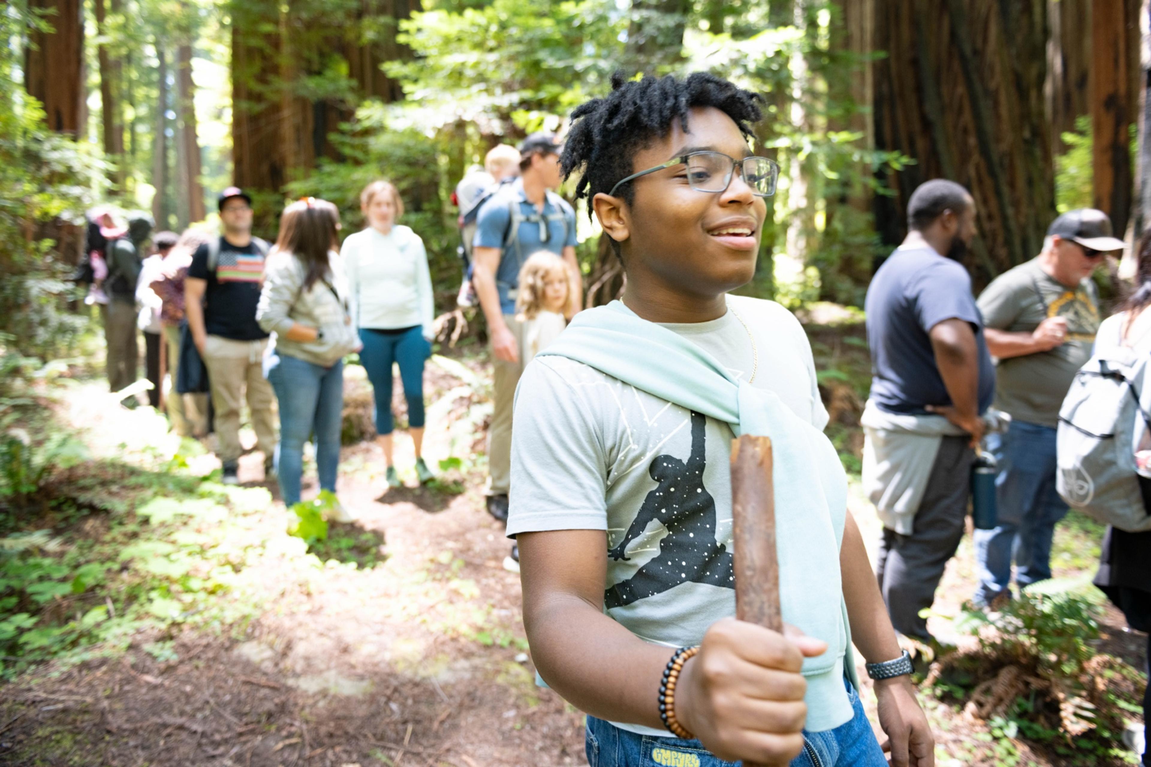 A teen holding a wooden walking stick at the front of a long line of hikers on a redwood trail A teen holding a wooden walking stick at the front of a long line of hikers on a redwood trail