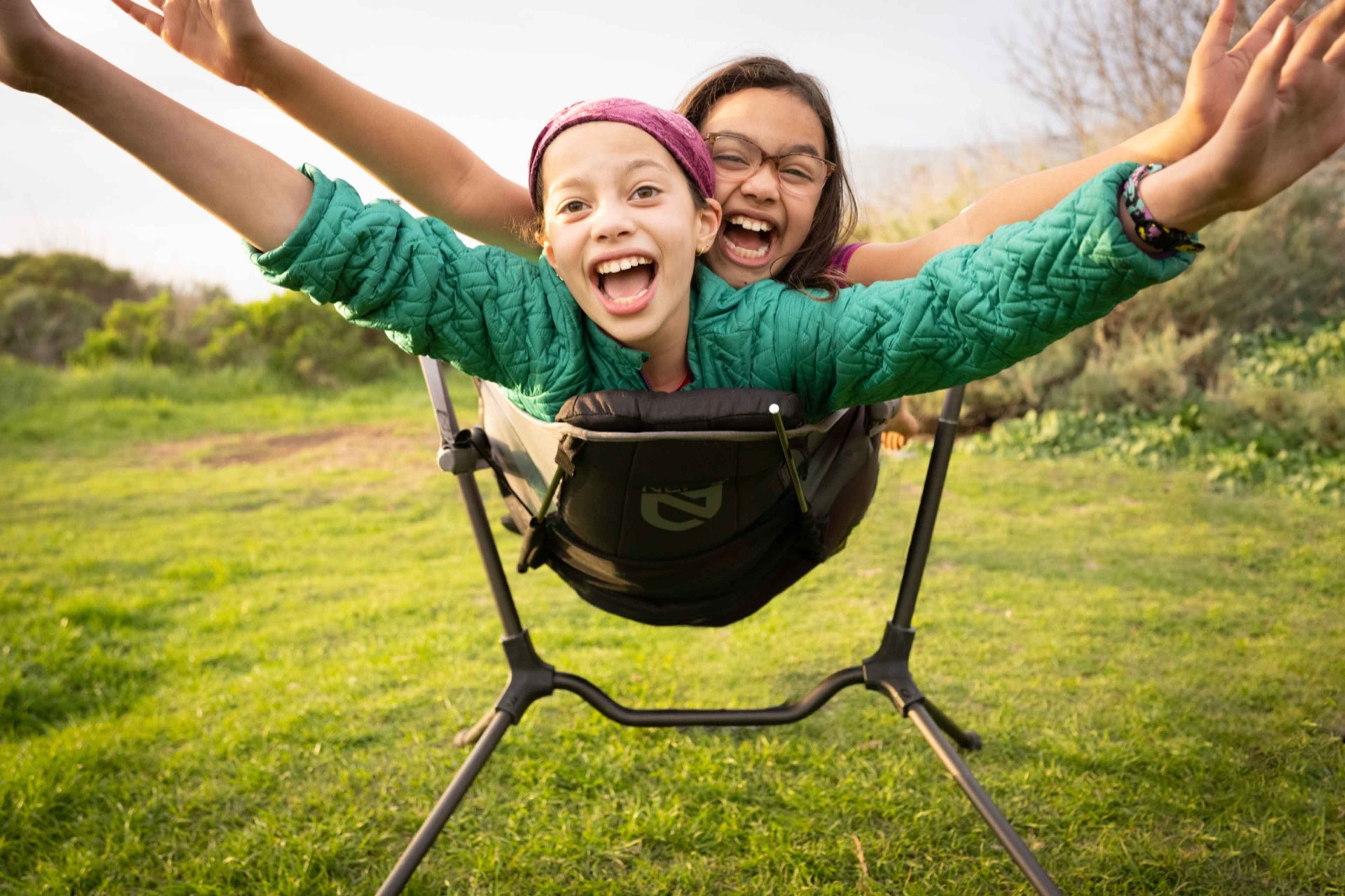 Two girls laughing with arms spread wide in a hammock chair on green grass at Kirk Creek Campground on the Big Sur coast