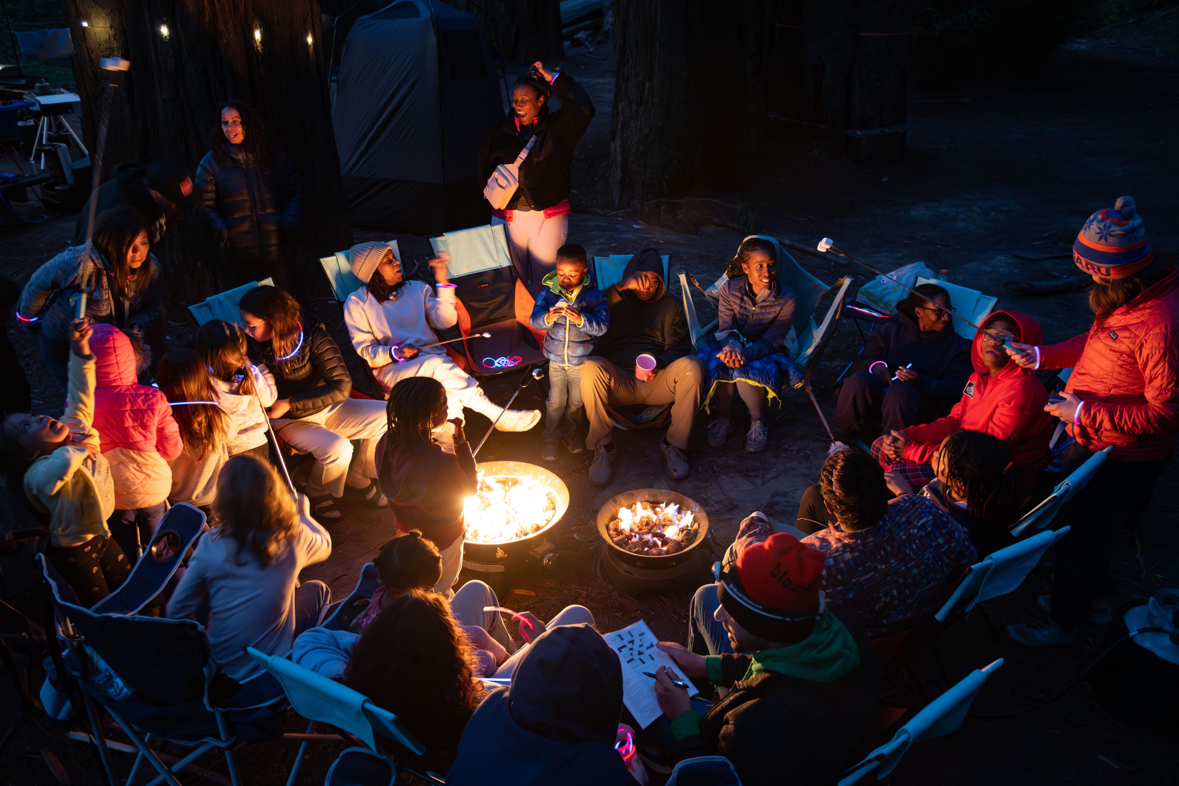 Roughly 30 people from a group camping trip seated in folding chairs around two Outland Living propane fire pits at Williams Grove, with kids holding marshmallow sticks and string lights overhead Roughly 30 people from a group camping trip seated in folding chairs around two Outland Living propane fire pits at Williams Grove, with kids holding marshmallow sticks and string lights overhead