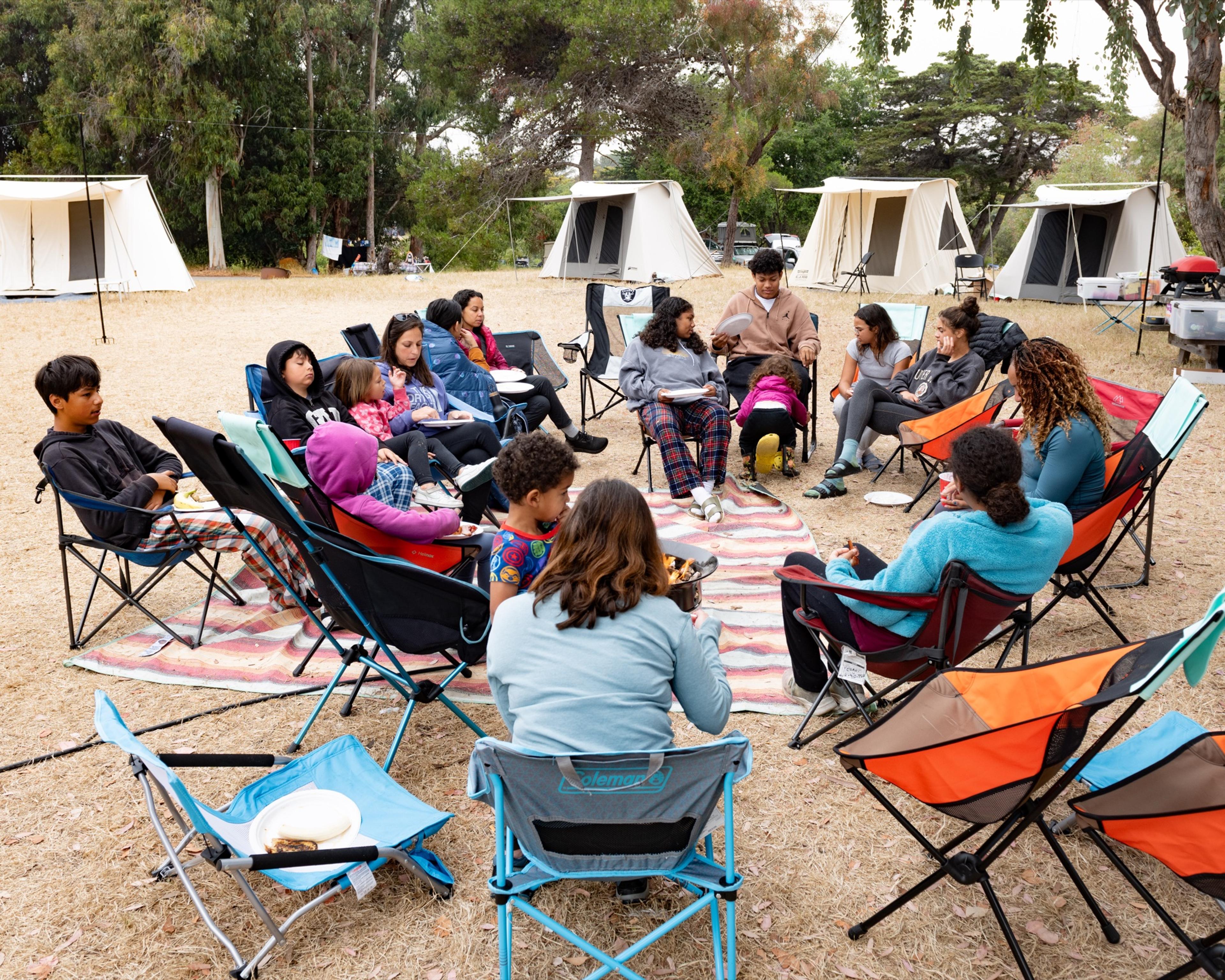 Adults and kids in a circle of folding camp chairs set on a striped woven rug around a small fire bowl, four canvas tents behind on dry grass Adults and kids in a circle of folding camp chairs set on a striped woven rug around a small fire bowl, four canvas tents behind on dry grass