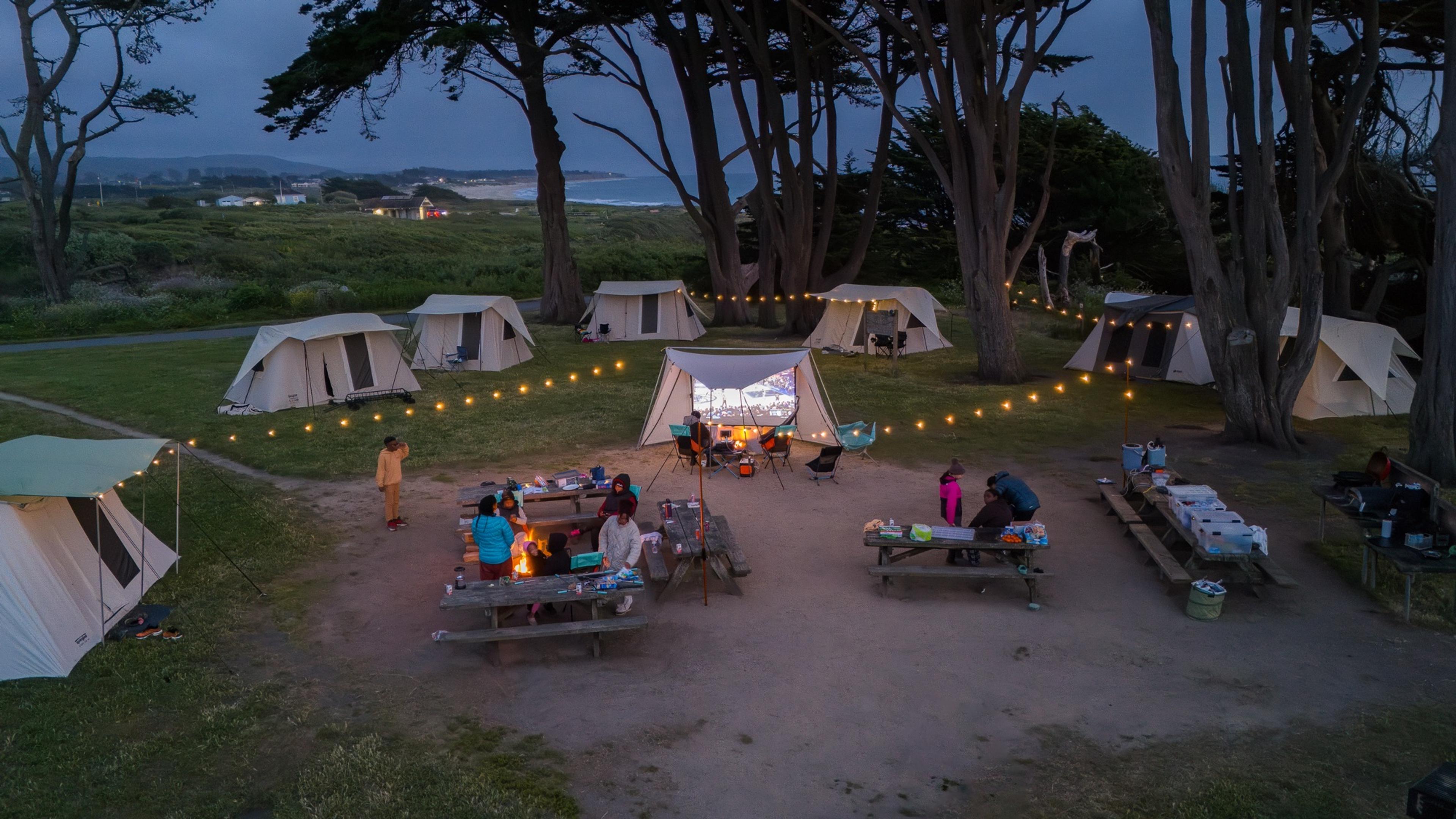 Drone view at dusk of a ring of canvas tents under cypress trees, string lights between them, and a movie playing on a screen in the middle of the group site, the ocean behind Drone view at dusk of a ring of canvas tents under cypress trees, string lights between them, and a movie playing on a screen in the middle of the group site, the ocean behind