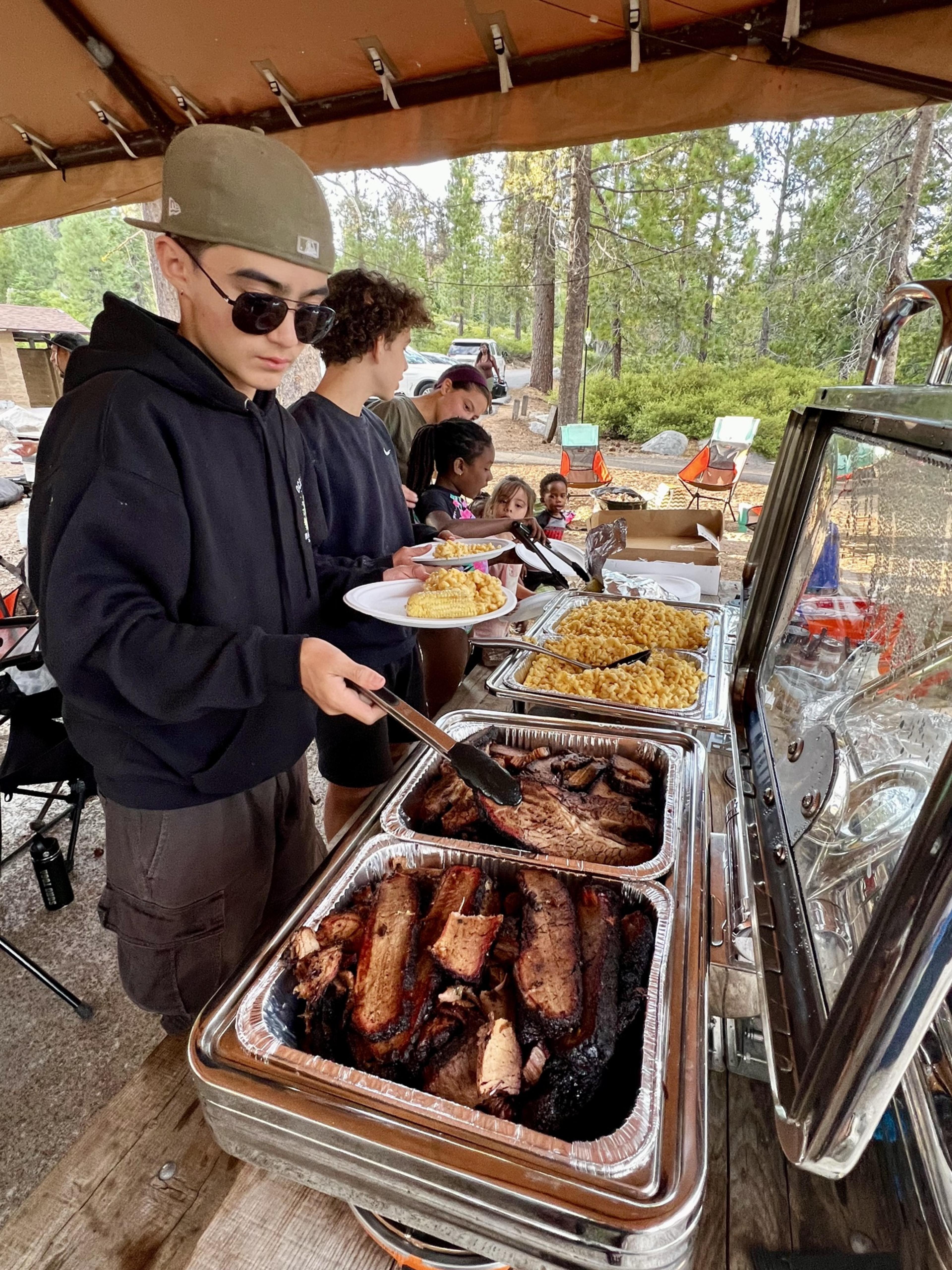 Serving line at the group camp, kids filling plates from open trays of barbecue and mac and cheese under the canopy Serving line at the group camp, kids filling plates from open trays of barbecue and mac and cheese under the canopy