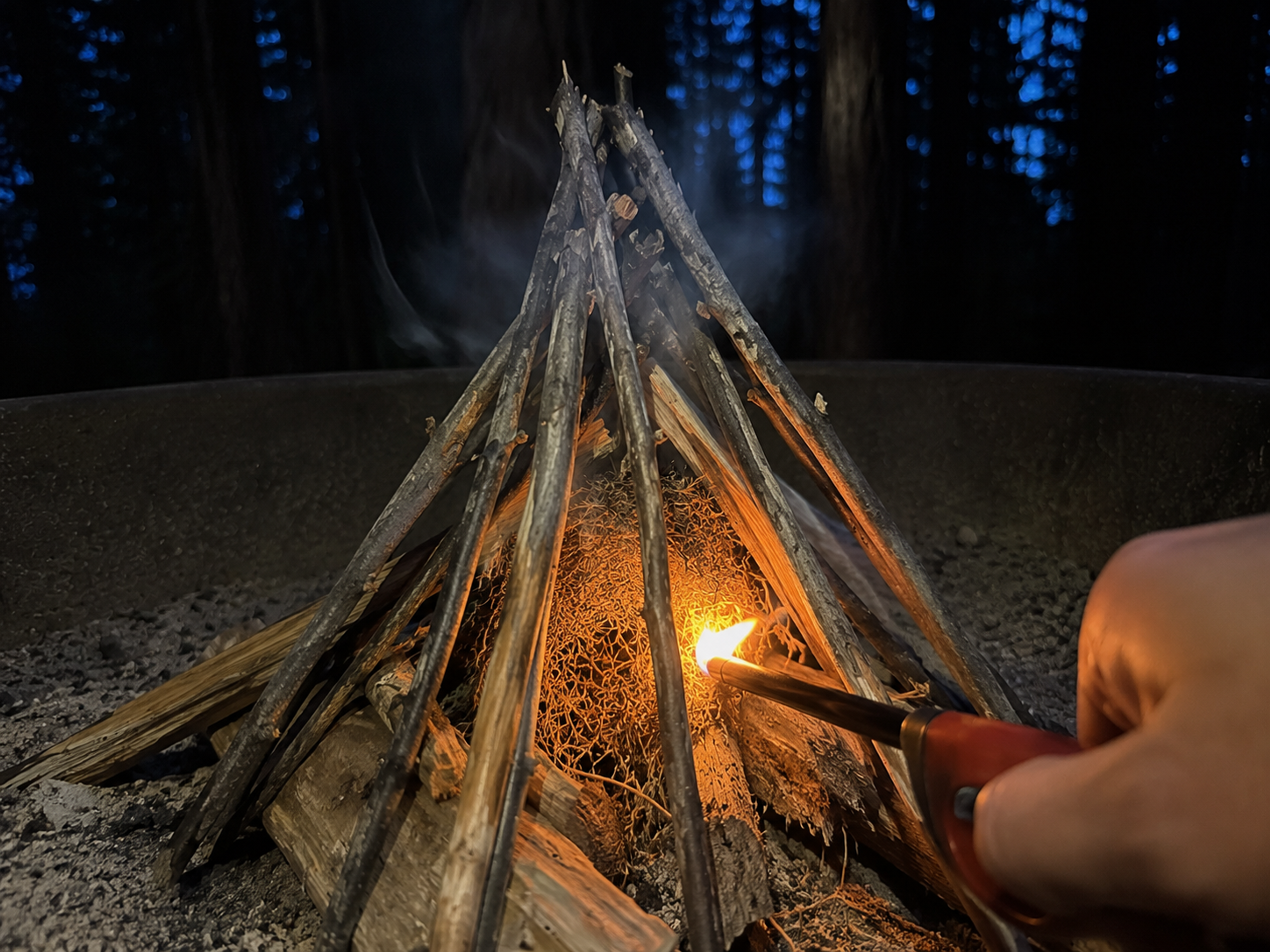 Close-up of a long-neck refillable lighter touching a pile of dry tinder at the base of an unlit teepee campfire, with the first small orange flame just catching, kindling sticks above not yet burning, dark trees in the background Close-up of a long-neck refillable lighter touching a pile of dry tinder at the base of an unlit teepee campfire, with the first small orange flame just catching, kindling sticks above not yet burning, dark trees in the background