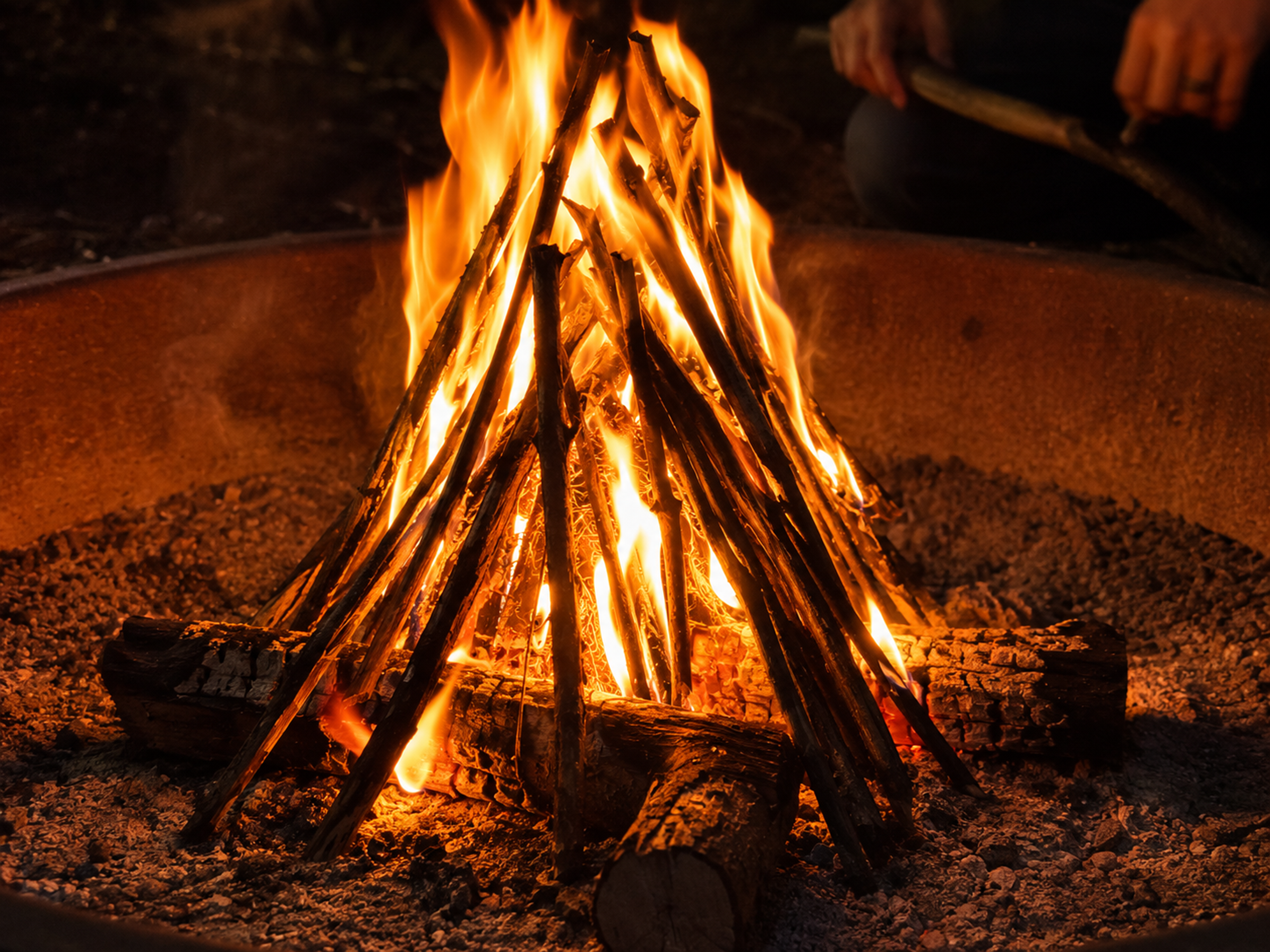 A well-built campfire burning brightly mid-burn with orange flames climbing through a teepee of kindling with visible air gaps between the sticks, two larger logs catching fire at the edges, minimal smoke, hands of an adult holding a stick visible in the blurred background A well-built campfire burning brightly mid-burn with orange flames climbing through a teepee of kindling with visible air gaps between the sticks, two larger logs catching fire at the edges, minimal smoke, hands of an adult holding a stick visible in the blurred background