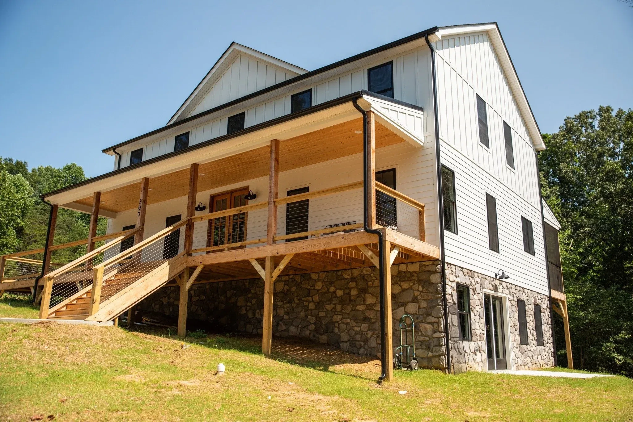 A multi-story house with white siding and a wooden deck on a grassy hill, surrounded by trees.