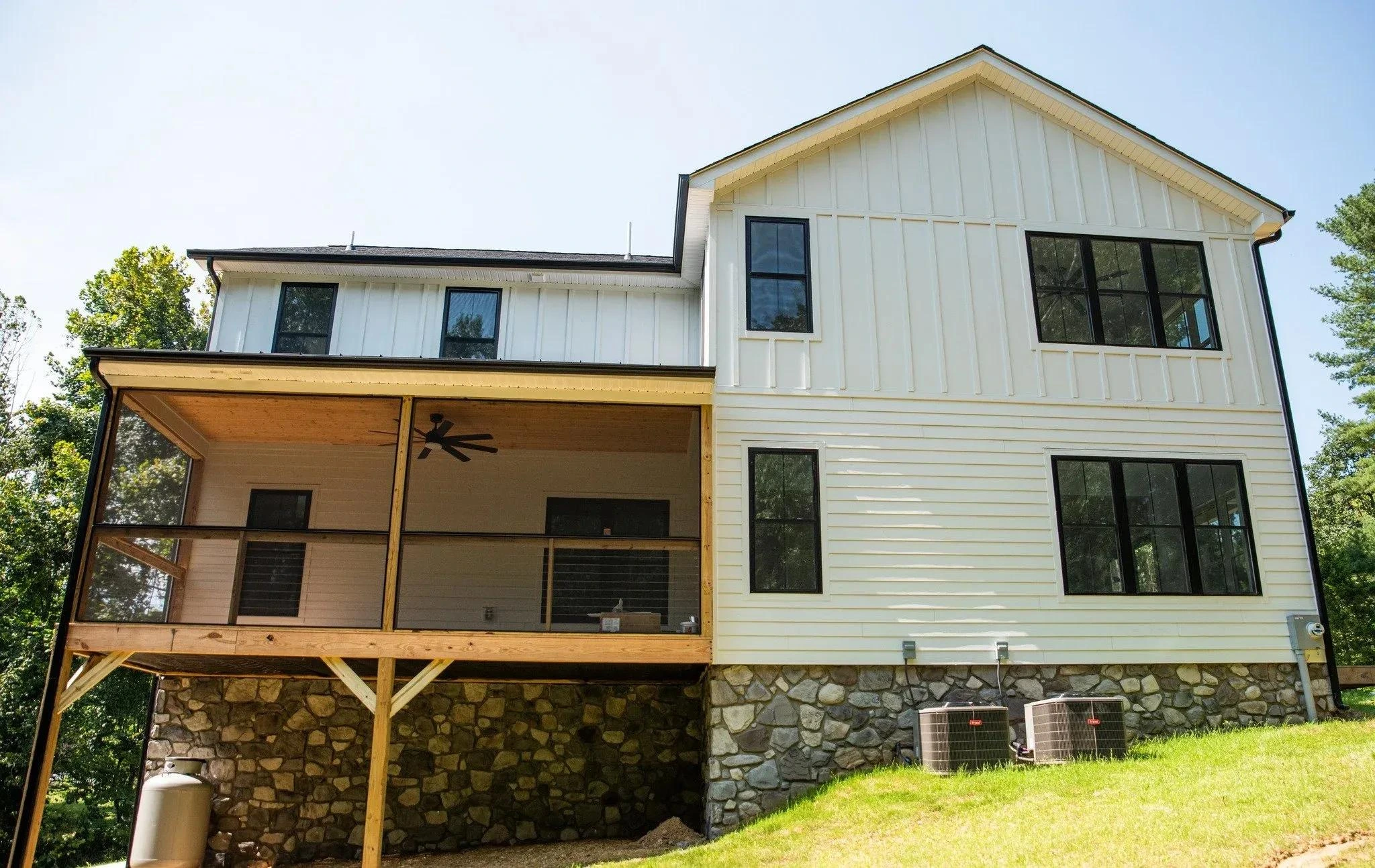 Modern two-story house with a stone foundation, white vertical siding, black window frames, and a screened-in porch with a ceiling fan.