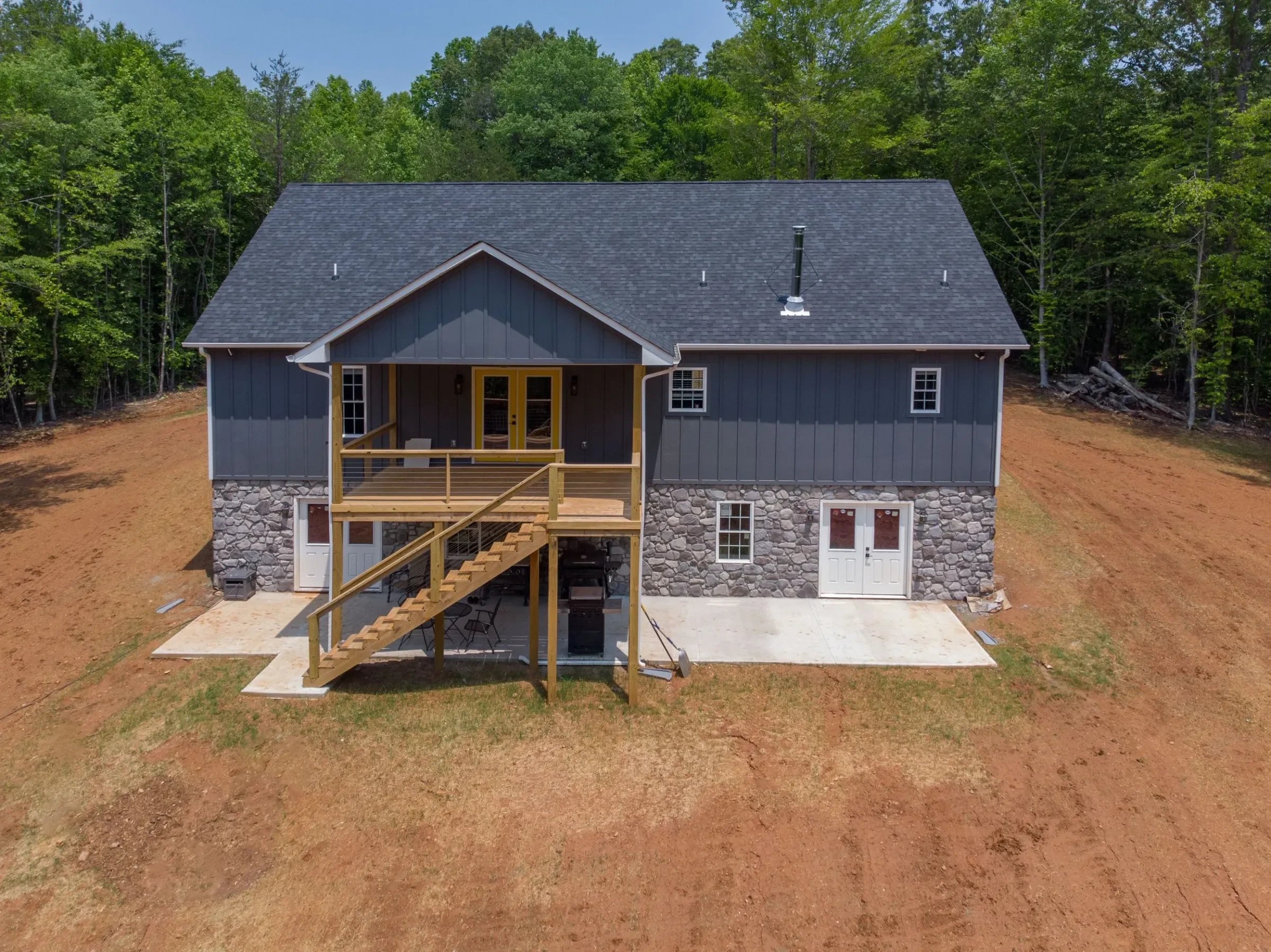 Back of a newly built house with blue siding, a stone foundation, and a wooden deck, with woods behind it.