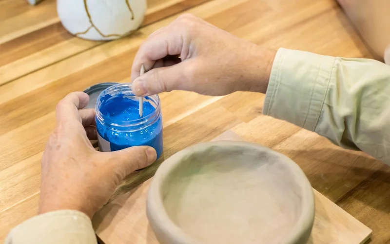 Manos mezclando pintura azul en un recipiente, junto a un cuenco de cerámica sin terminar.