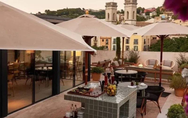 Terraza del Hotel Casa Cacao con mesas, sillas y sombrillas beige, bar con botellas y flores púrpuras ante vistas de Girona al atardecer