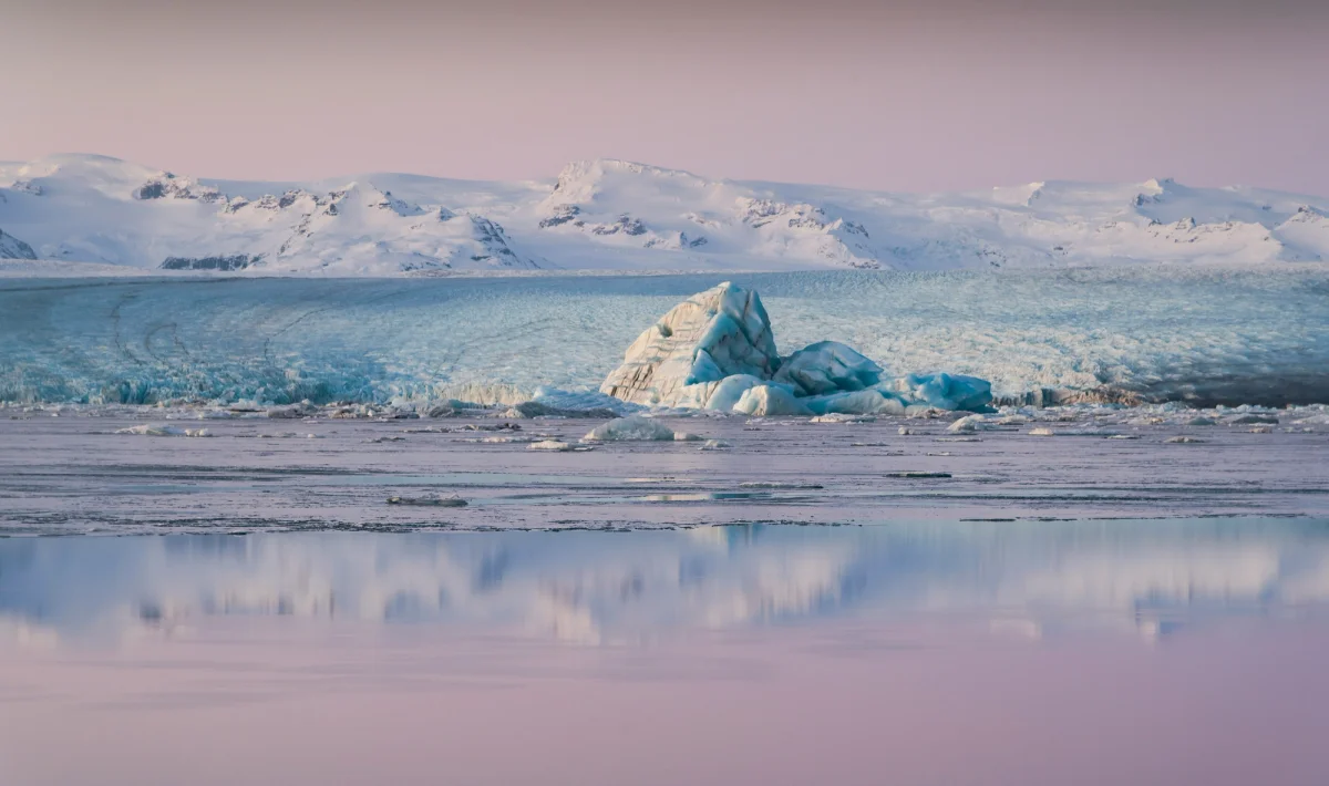 Glaciar azul y blanco con montañas nevadas al fondo, reflejado en agua tranquila.