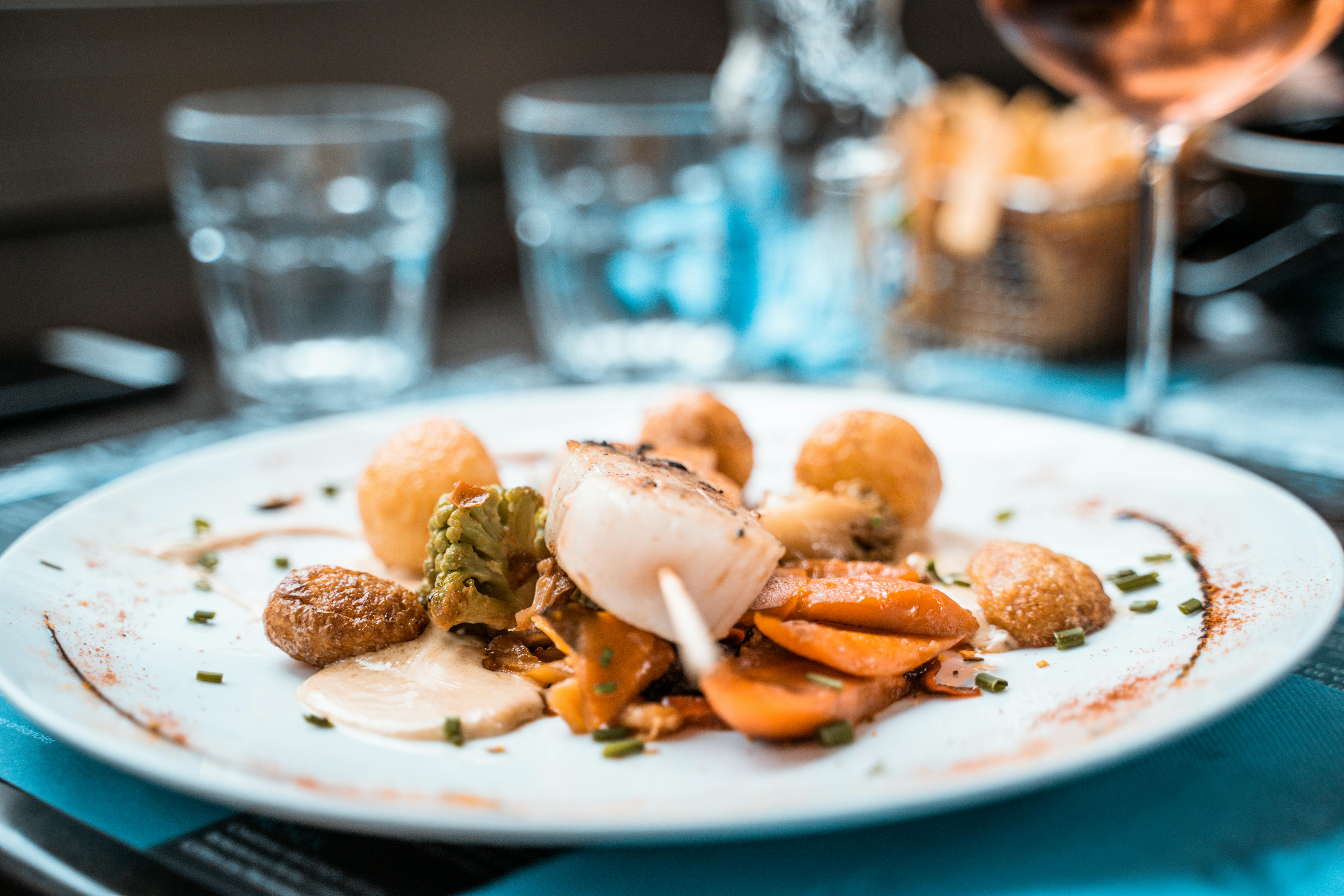 Plato gourmet con vieira, verduras, patatas y salsa en una mesa con copas de fondo.