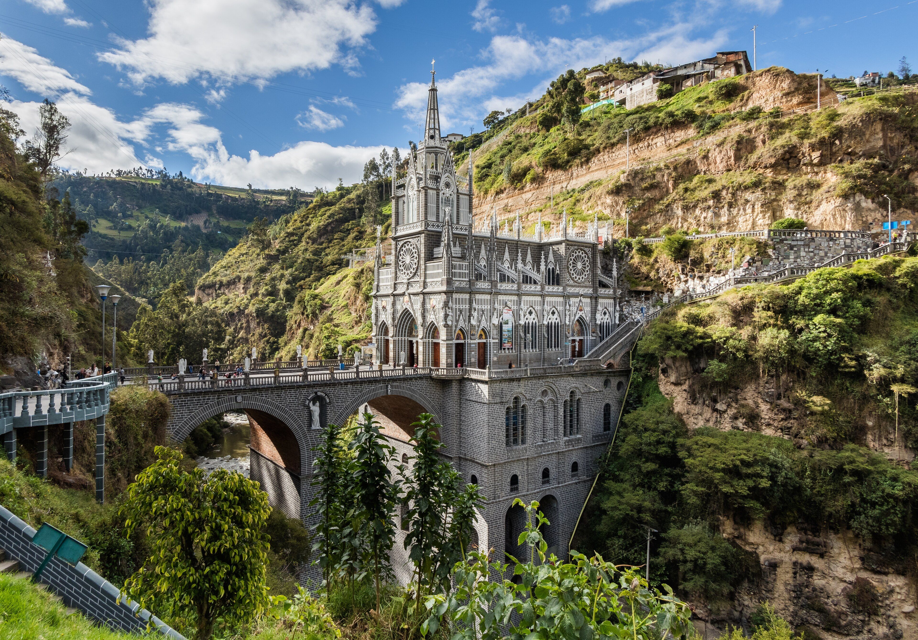 Iglesia neogótica gris con torre puntiaguda sobre un puente de piedra de arcos que cruza un cañón verde, bajo un cielo azul con nubes.