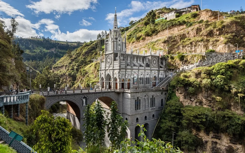 Iglesia neogótica gris con torre puntiaguda sobre un puente de piedra de arcos que cruza un cañón verde, bajo un cielo azul con nubes.