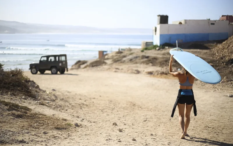 Mujer caminando por la playa de Agadir con tabla de surf azul bajo el brazo, jeep negro al fondo y olas del mar Atlántico