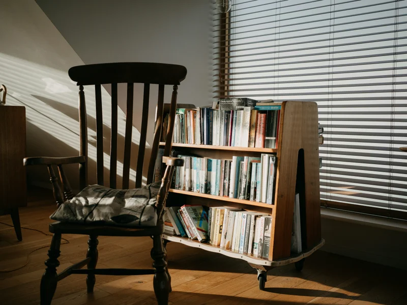 Una silla de madera y una estantería con libros junto a una ventana con persianas.