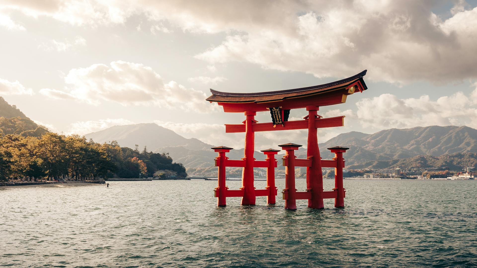 El torii rojo del santuario Itsukushima emerge del mar en Miyajima, con montañas y follaje otoñal al fondo