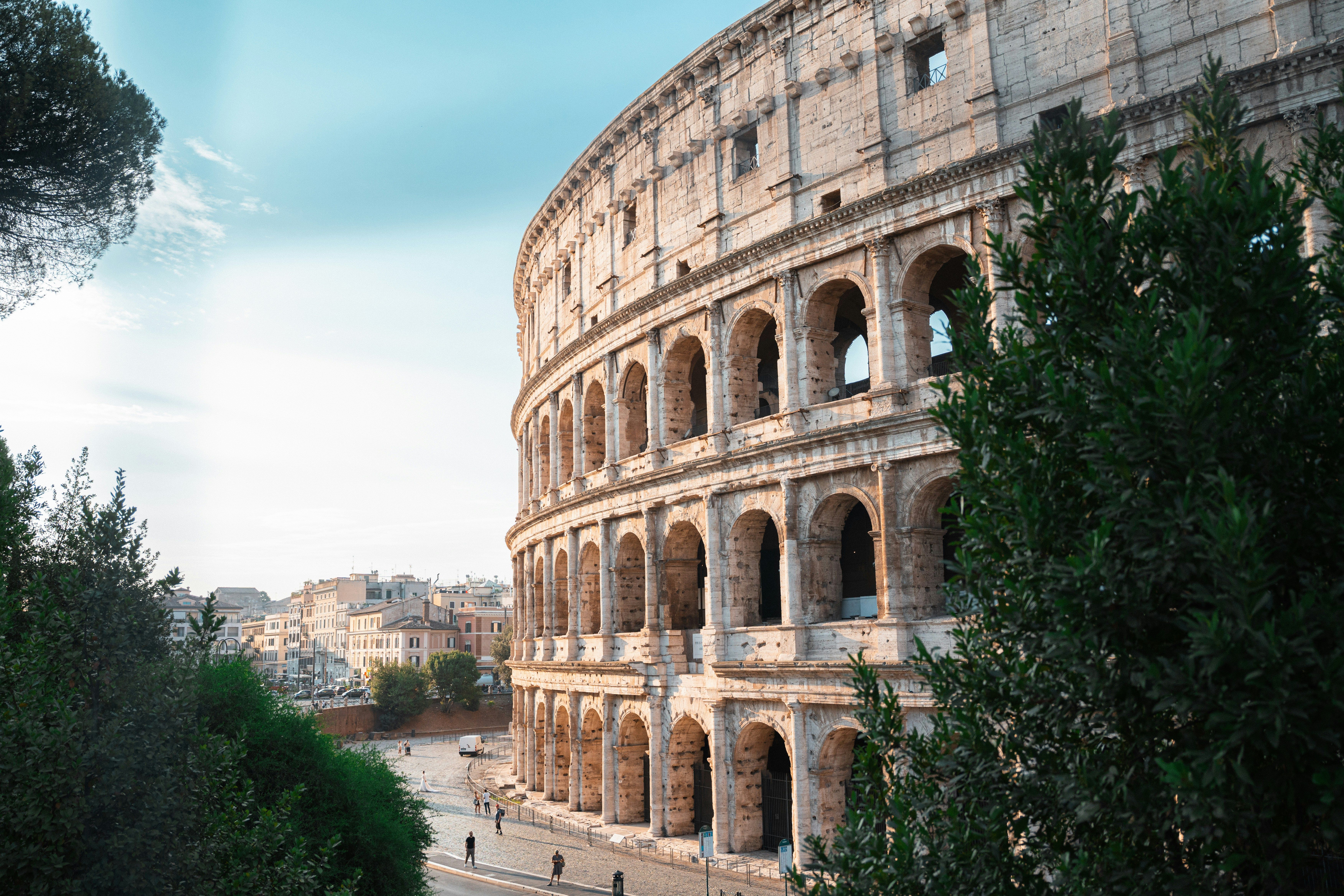 Vista parcial del Coliseo romano con arcos y columnas, rodeado de vegetación y cielo azul.