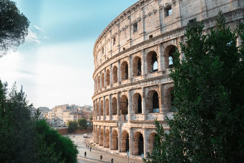Vista parcial del Coliseo romano con arcos y columnas, rodeado de vegetación y cielo azul.