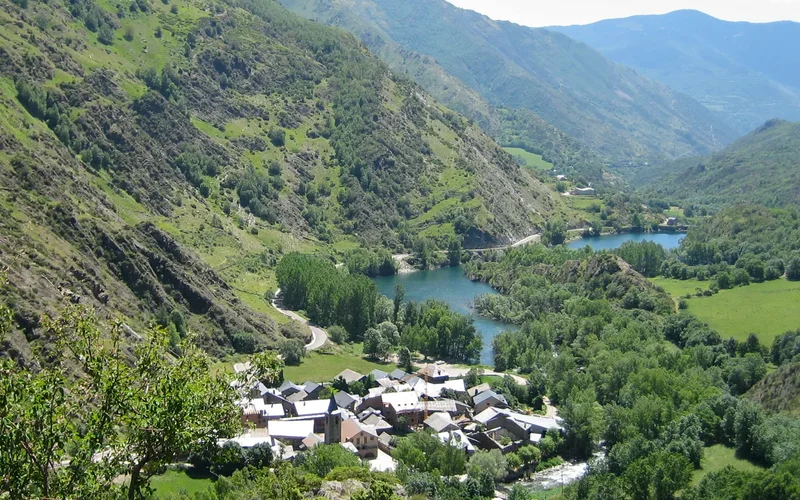 Vista panorámica de un valle montañoso con prados verdes, un pueblo de casas tradicionales y un lago sereno entre montañas en Pallars Sobirá