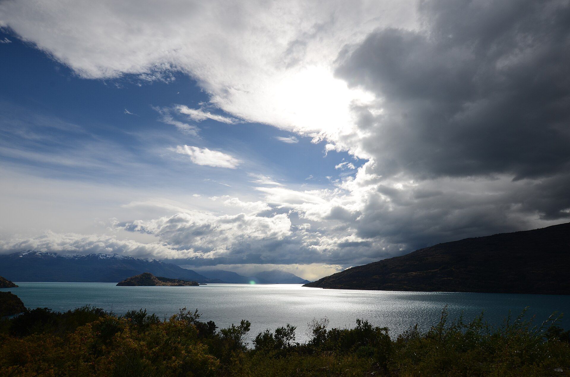 Vista panorámica de un lago turquesa bajo un cielo nublado con montañas al fondo.